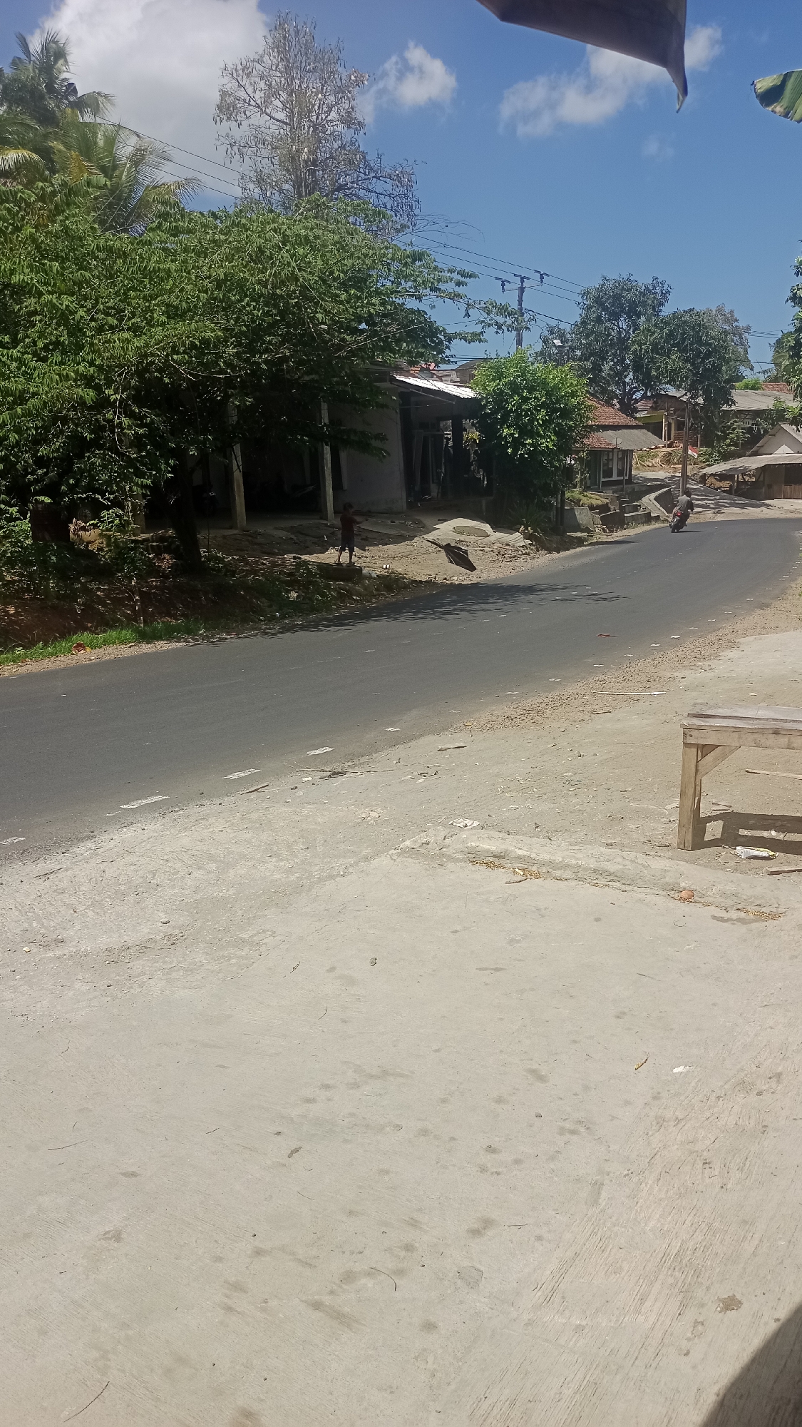 The image depicts a street scene in Cikalong, Indonesia, under a bright blue sky with scattered white clouds. The foreground is dominated by a light-colored concrete surface with some dirt and debris. To the right, a simple wooden bench with two legs is visible. A paved asphalt road runs diagonally from the bottom left to the upper right of the frame. Along the left side of the road, there is a grassy embankment with trees and vegetation. On the right side of the road, several houses with tiled roofs and open-air structures are visible, nestled among trees. In the middle ground, a person, appearing to be a child, stands on the embankment near a house. Further down the road, a motorcycle is being ridden. The overall environment suggests a rural or semi-urban setting in Indonesia.