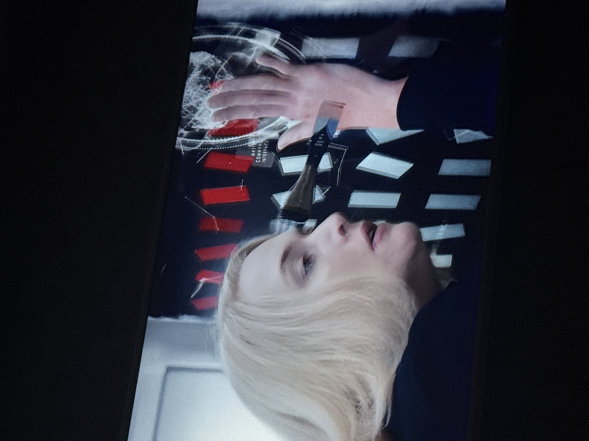 A screen displays a woman with blonde hair, facing upwards and slightly to the right, her mouth slightly open. Her right hand is visible, palm down, against a backdrop of red and white rectangular shapes. Above her hand, there are translucent graphics resembling a web or circular patterns. A dark-colored lever or handle is positioned between her face and hand. Text reading "MASTER COMMAND INTERFACE" is visible. No location information is provided.