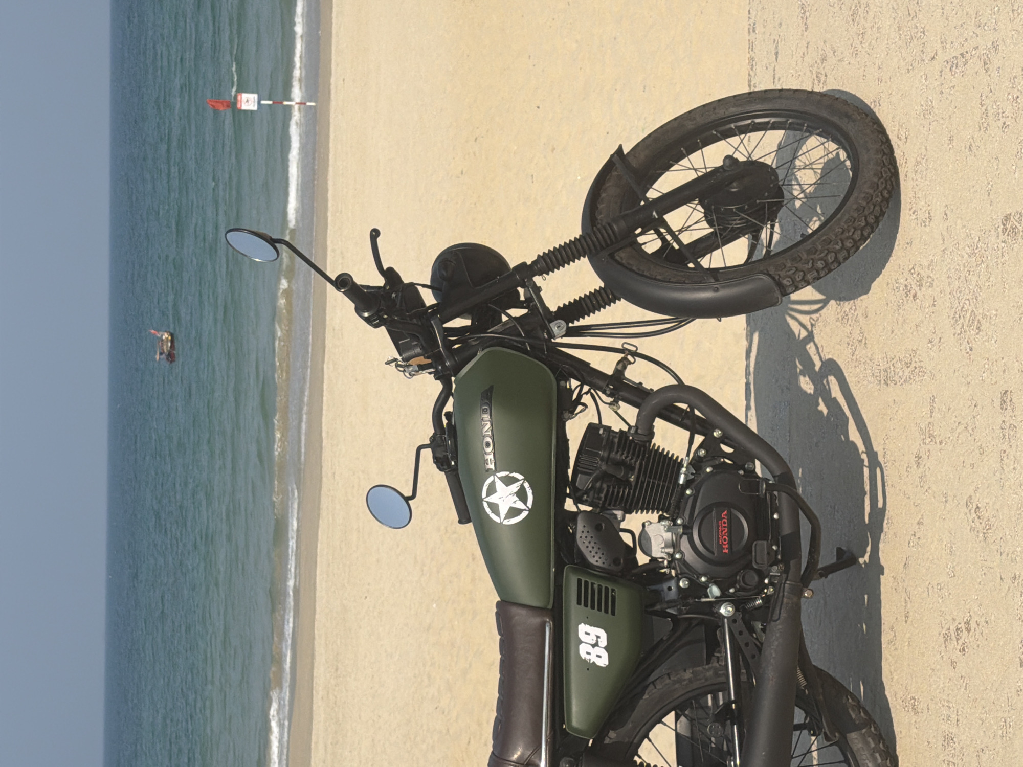 The image depicts a green Honda motorcycle parked on a sandy beach. The beach is adjacent to a body of water, likely the ocean, which is visible in the background. A red and white safety flag stands near the water's edge, and a person can be seen in the distance. The motorcycle has a white star decal on the gas tank and the number "89" painted on the side panel. The motorcycle appears to be photographed in Đà Nẵng, Vietnam based on its location on the coast of the sea.