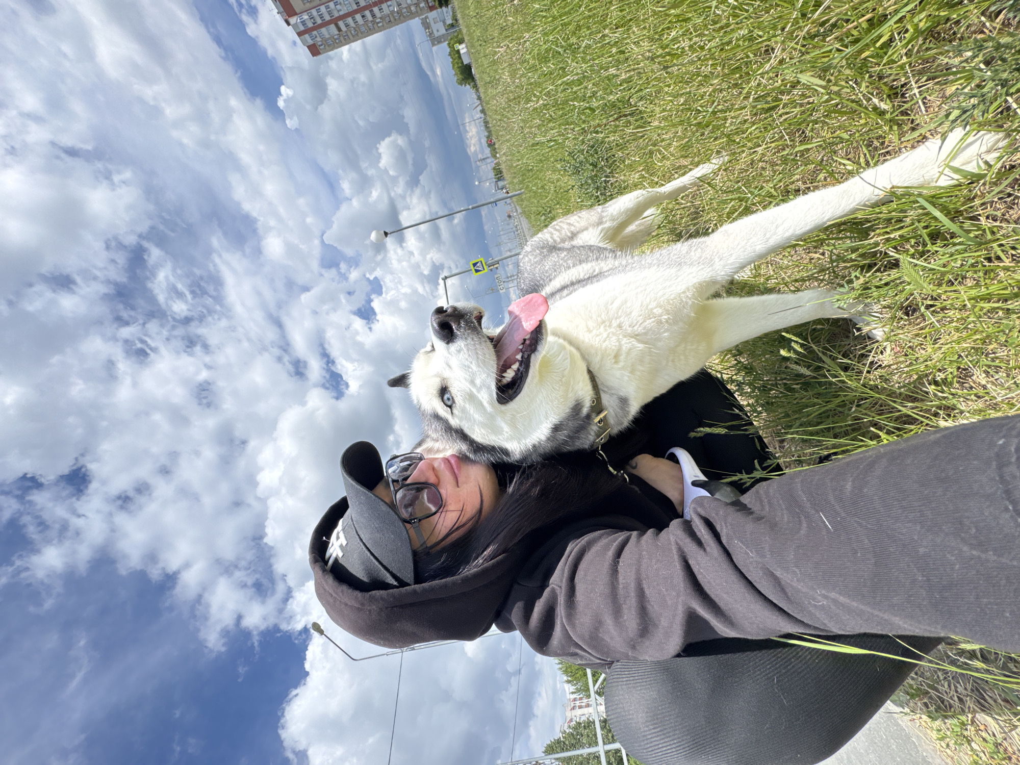 The image shows a selfie of a person with long black hair wearing a black hooded sweatshirt, a black baseball cap, and sunglasses. The person is positioned at the bottom center of the frame, looking up at the camera. Behind them is a white and grey Siberian Husky dog with its mouth open and tongue out, appearing to be panting or smiling.
The background is composed of a partly cloudy sky with blue patches visible among the clouds, and a grassy area with a sidewalk to the right. A street sign and lamp post are seen in the distance. In the upper part of the image, there is a multi-story building.
The location appears to be an urban park or green space. It is located in the city of Unknown, Unknown. The scene is taken during daylight hours.