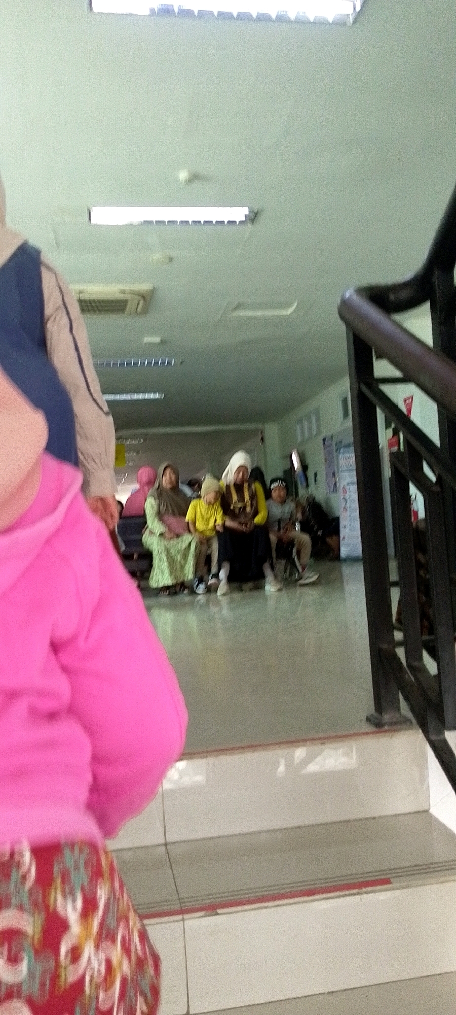 The image captures an indoor scene, likely a waiting area, with a group of people seated and standing. In the foreground, a person wearing a bright pink hooded jacket and a patterned skirt is partially visible, facing away from the camera. Behind them, a railing of a staircase is on the right side of the frame.
Further back, a group of individuals are seated. One woman wears a green patterned dress and a headscarf. Next to her is a child in a yellow outfit and a beige hat. To their right, another woman is seated, wearing a yellow top and a white headscarf, with a child next to her wearing a black and white striped hat. Another individual in grey is seated to the far right. They appear to be in a well-lit room with fluorescent lighting panels on the ceiling and an air conditioning unit visible. Posters and a white banner are present on the wall in the background. The floor is a light gray, possibly tiled.
This scene is situated within the city of Sumenep, Indonesia. The attire of the individuals, particularly the headscarves and modest clothing, suggests a cultural context common in Indonesia. The overall atmosphere appears calm, possibly indicating a clinic, government office, or public service center.