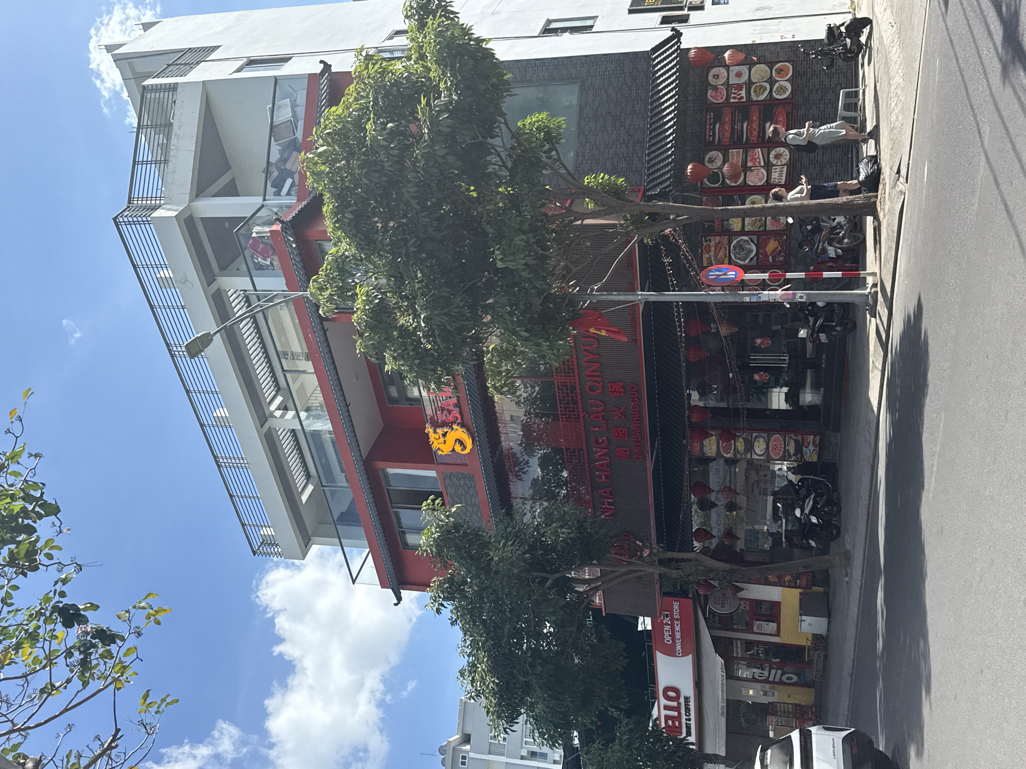 The scene depicts a street in Nha Trang Ward, Vietnam, featuring a multi-story building with a distinct architectural style, primarily white with red and grey accents. The building appears to house a restaurant named "Nha Hang Lau Qinyu," indicated by signage in Vietnamese and Chinese characters. A golden dragon ornament is also visible on the building's facade.
Several trees line the street, partially obscuring the view of the building. On the sidewalk, individuals are seen either sitting or lying down. Parked motorcycles are also present on the sidewalk.
The scene also includes a convenience store named "Hello," with its signage prominently displayed. Traffic signs are visible near the restaurant. The sky is partly cloudy.