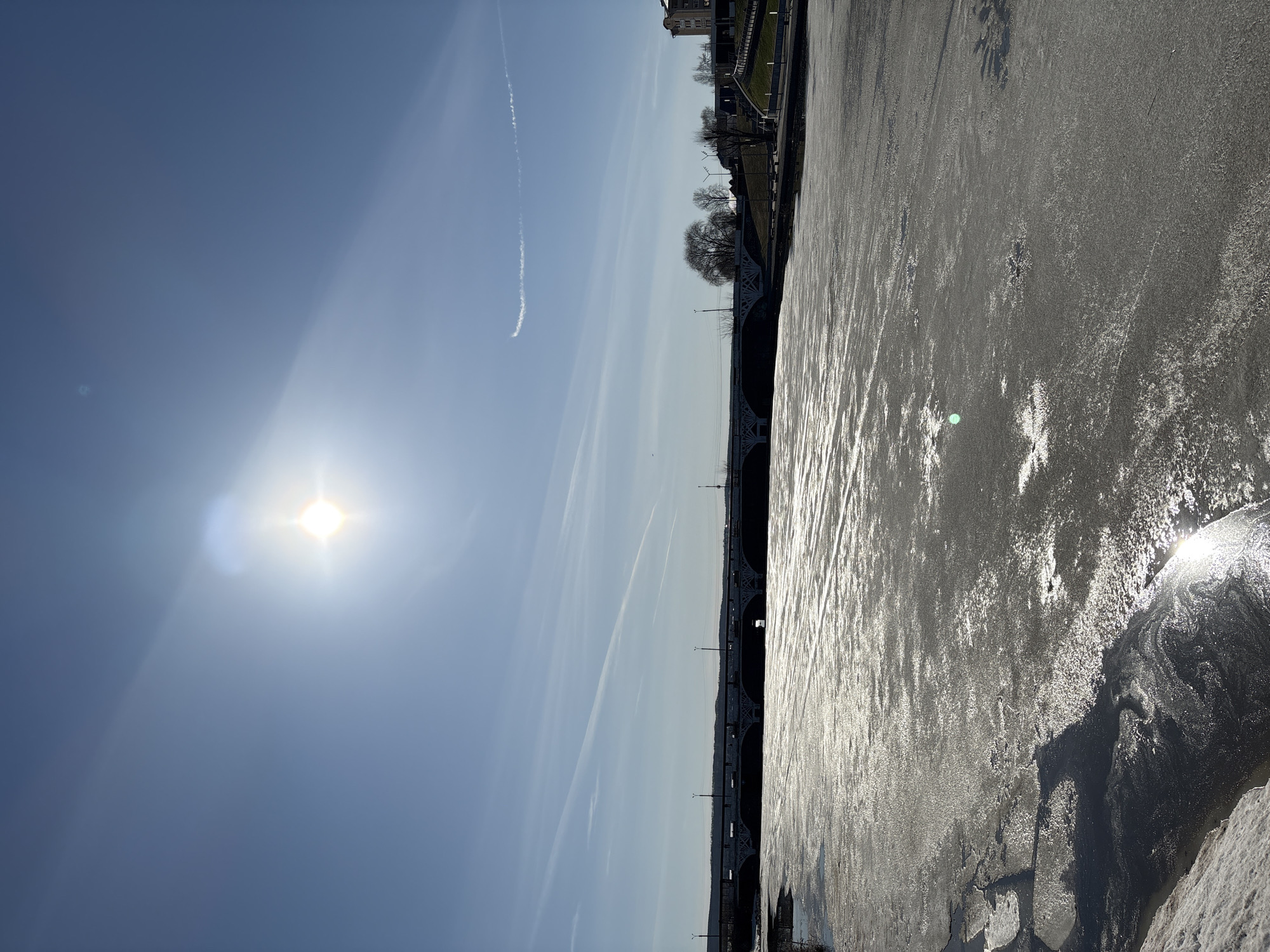 The image captures a broad landscape scene under a bright, clear blue sky. The photo is taken horizontally, with a vibrant sun positioned in the upper left quadrant, creating a strong lens flare and illuminating the scene. Several faint contrails from aircraft are visible across the sky, with one prominent, curving contrail stretching from the upper center towards the middle left.
In the midground, a large body of water, likely a river or lake, stretches across the frame. The water's surface appears partially frozen or covered in slushy ice, intensely reflecting the bright sunlight, which creates a shimmering, textured effect. A multi-arched bridge with intricate ironwork and streetlights spans the water in the middle distance, connecting the near and far banks.
On the distant bank, a shoreline rises, featuring bare trees suggesting a winter or early spring setting, and several buildings on a slightly elevated terrain. The sun's angle indicates it is daytime, possibly late morning or early afternoon. The overall impression is one of a crisp, bright day, likely cold given the state of the water. There is no visible text in the image. The location is Votkinsk, Russia.