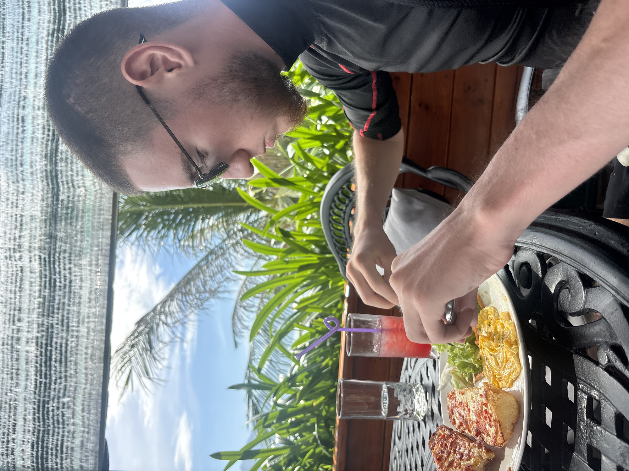 A young man with short brown hair and glasses is eating breakfast at a table outdoors. He is wearing a black shirt with red stripes. On the table is a plate of food with two pieces of toast with melted cheese, a salad, and scrambled eggs. There are also two drinks, one with a purple straw, and a glass of water. In the background, there are palm trees and the sky. The scene appears to be a cafe or restaurant in Bắc Nha Trang, Vietnam.