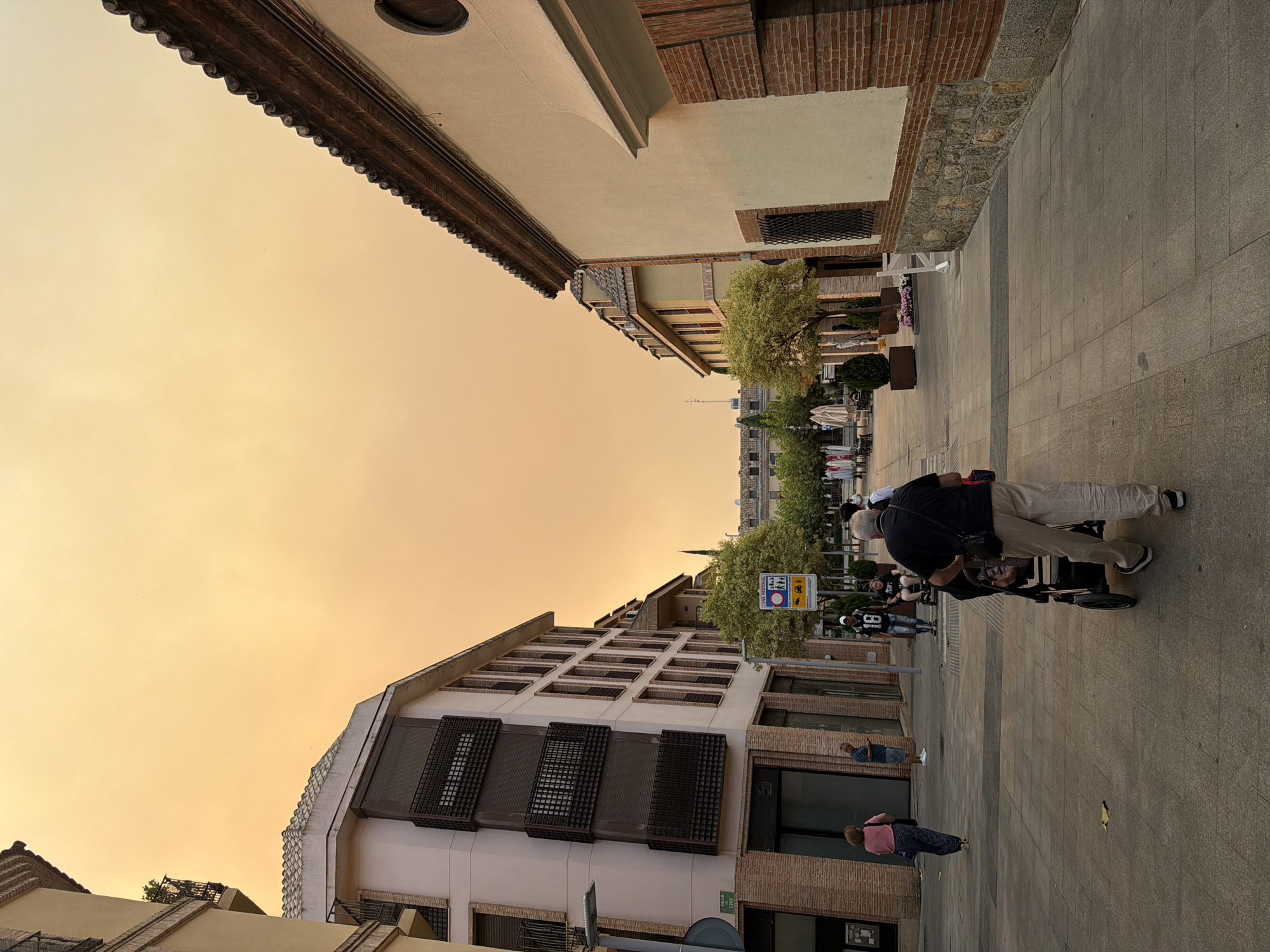 In Las Rozas de Madrid, Spain, on a pedestrian street, an orange haze fills the sky, casting an unusual light on the scene. Buildings line both sides of the street, with architecture featuring balconies and tiled roofs. The street is paved with large stone tiles. Several people are present. In the foreground, a person walks away from the camera, pushing a stroller. Further down the street, other individuals are walking or standing, some near trees and outdoor cafe seating. A street sign indicating a pedestrian zone is visible. The overall atmosphere is subdued due to the atmospheric conditions.