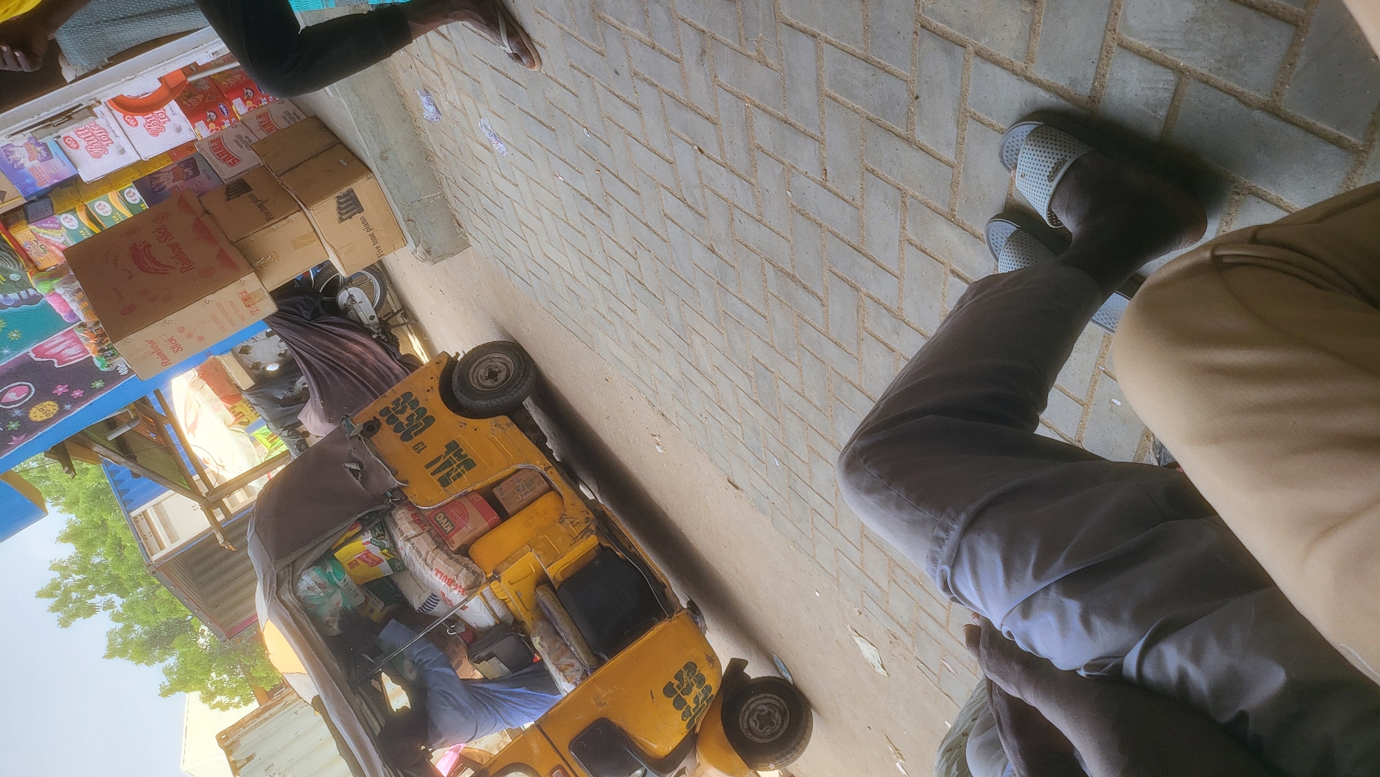 In Fagge C, Nigeria, a yellow auto-rickshaw, laden with various goods, is parked on a paved surface. The rickshaw has the license plate "MAL 19 0338". A person wearing a blue shirt and grey pants is partially visible inside the rickshaw, appearing to be engaged in loading or unloading. In the foreground, another individual, also wearing grey pants and sandals, is seated, with their legs crossed. Behind the rickshaw, stacks of boxes are visible, suggesting a market or distribution point. The environment appears to be an outdoor commercial area with trees and some structures in the background.