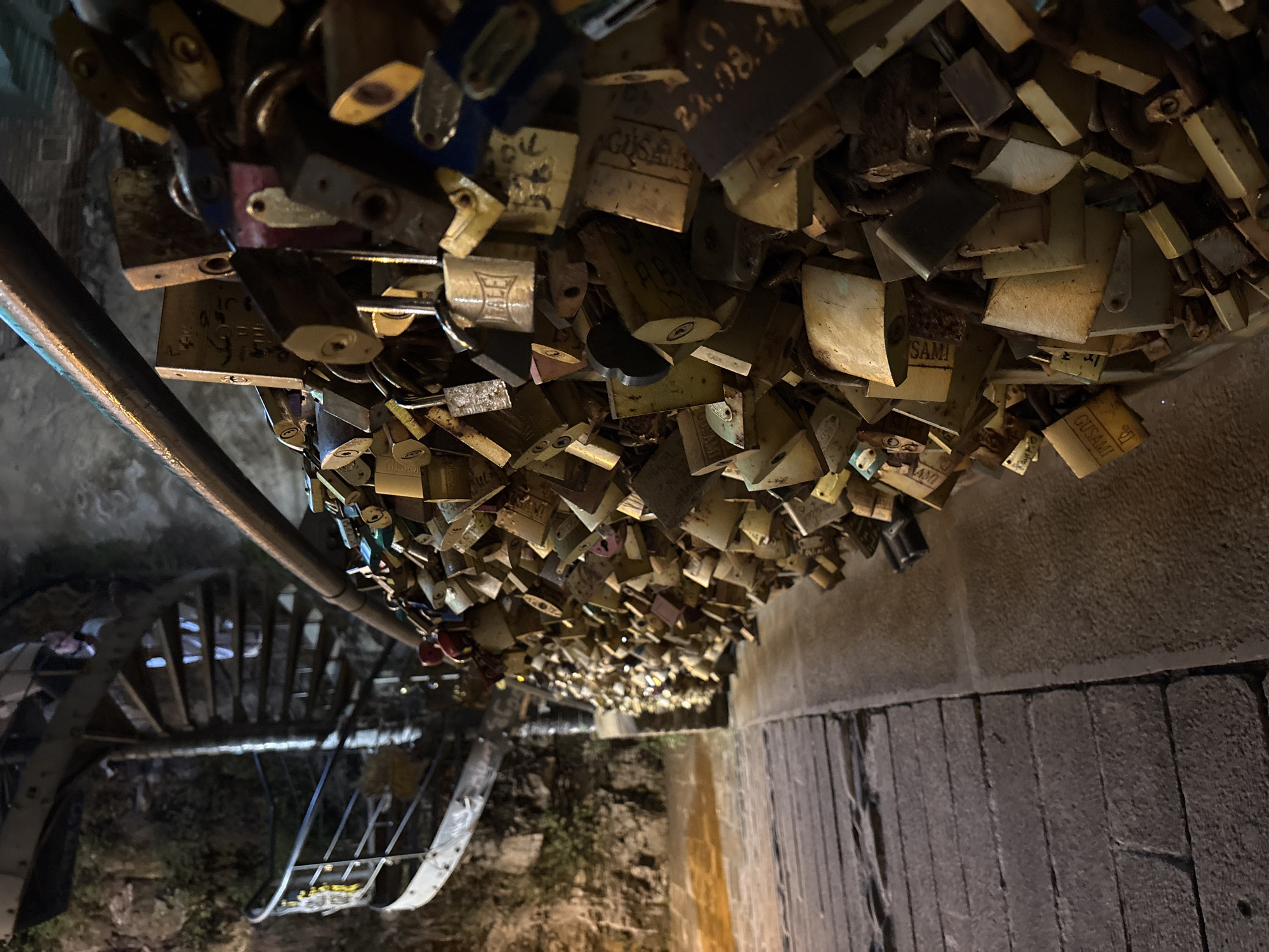 The image depicts a large collection of padlocks, many of which appear to be love locks, attached to a railing or fence. The padlocks are of various sizes, shapes, and colors, with some showing signs of rust and age. Many are inscribed with names, initials, dates, or short messages, though the text is often difficult to read due to the angle, lighting, and the sheer density of the locks.
The scene is viewed from an elevated perspective, looking down and across. To the left, a metal railing runs diagonally across the frame, with a spiral staircase visible beneath it. The overall setting appears to be an outdoor location, possibly a bridge or a walkway, given the presence of the railing and the stone-like structure on the right side of the image.
The time of day is suggested to be evening or night by the dim lighting and the overall dark tone of the image. The lighting appears to be artificial, casting a warm glow on some of the locks and creating shadows. There are no visible people in the frame. The mood is one of quiet accumulation, with the locks representing countless shared moments or hopes. The sheer volume of locks suggests a place of significance for many people. The text on the locks, where legible, hints at personal inscriptions and dedications. For instance, some locks show inscriptions like "OPEN", "GUSAMI", and a date "22.08.14".