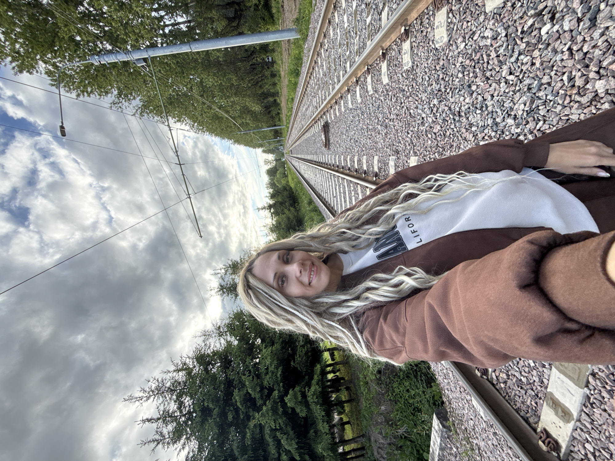 A woman with blonde dreadlocks is posing for a selfie along a railway track, which leads into the distance, lined with gravel and green vegetation. Overhanging trees and overhead power lines are visible along the track. The sky is overcast with gray clouds. The location is Rusanovo, Russia. The woman is wearing a brown jacket over a white t-shirt.