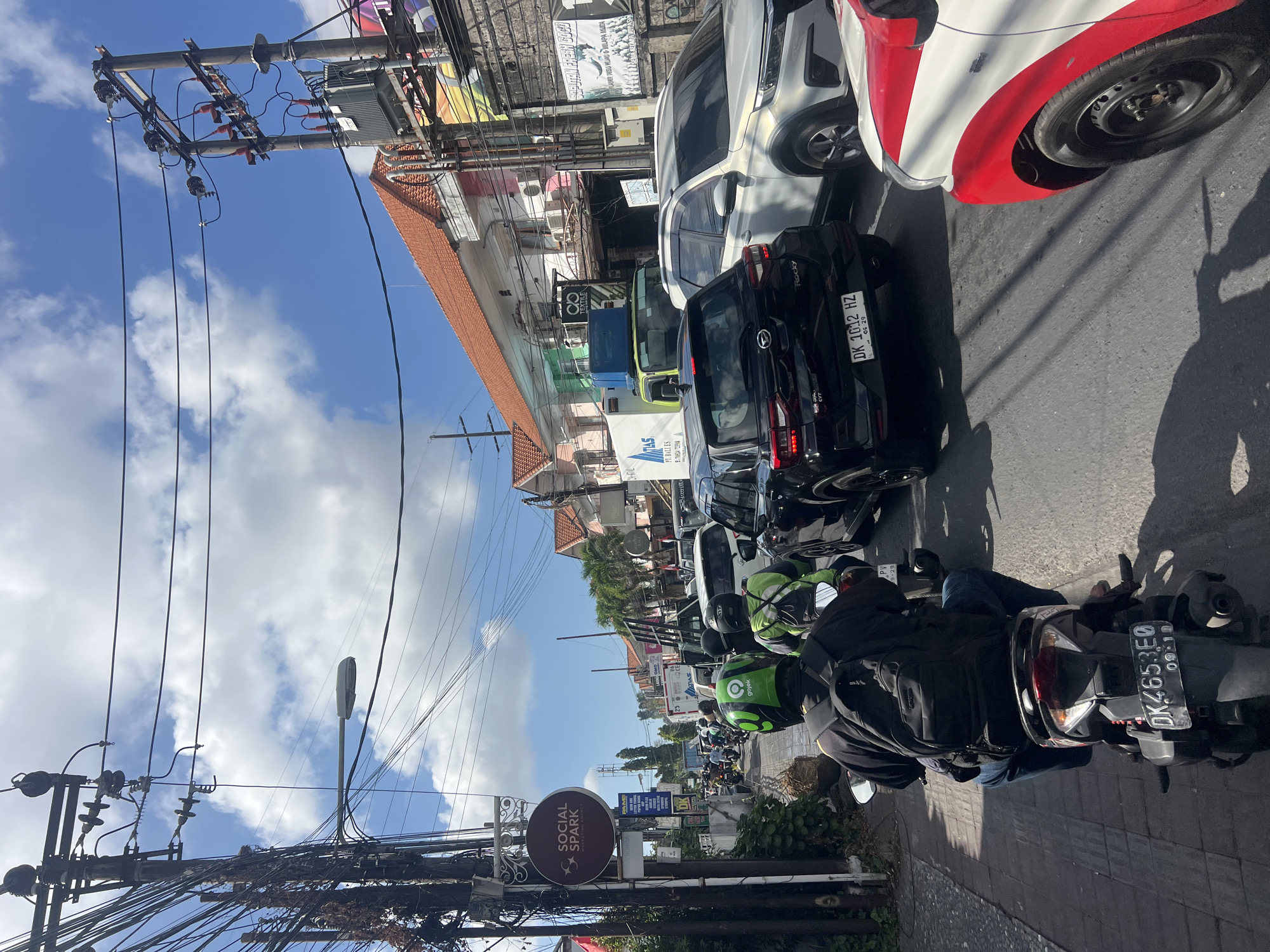 In Denpasar, Indonesia, a traffic jam is occurring on a street. A rider on a scooter, wearing a black backpack and blue jeans, is in the foreground, facing away from the camera. Other motorbikes with riders wearing helmets, some with green, are also visible in the traffic. Cars, including a black SUV with license plate "DK 1012 HZ" and a white SUV, are stuck in the congestion. Buildings line the street, featuring red-tiled roofs and storefronts with signage. Power lines and utility poles crisscross overhead, contributing to the urban landscape. The sky is blue with scattered white clouds.