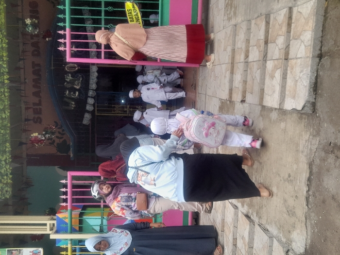 A group of people, including children in white uniforms, stand near a building with colorful gates and a "SELAMAT DATANG" sign. One woman is wearing a tan outfit, and others are dressed in black and blue. A child with a pink backpack walks on a sidewalk. The area appears to be the entrance to a school or community center, with stairs leading up to a platform.