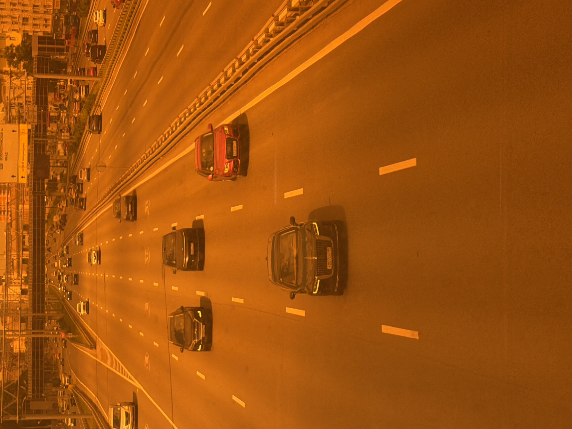 The image shows a highway with multiple lanes of traffic. Several cars are visible on the highway, moving in the same direction. The lighting suggests it is daytime, though the overall color cast of the image is orange, which might indicate a filter or atmospheric conditions. In the background, there are buildings and overpasses, indicating an urban or suburban environment. The road surface is asphalt, with clear lane markings. The perspective is from above, looking down on the highway.