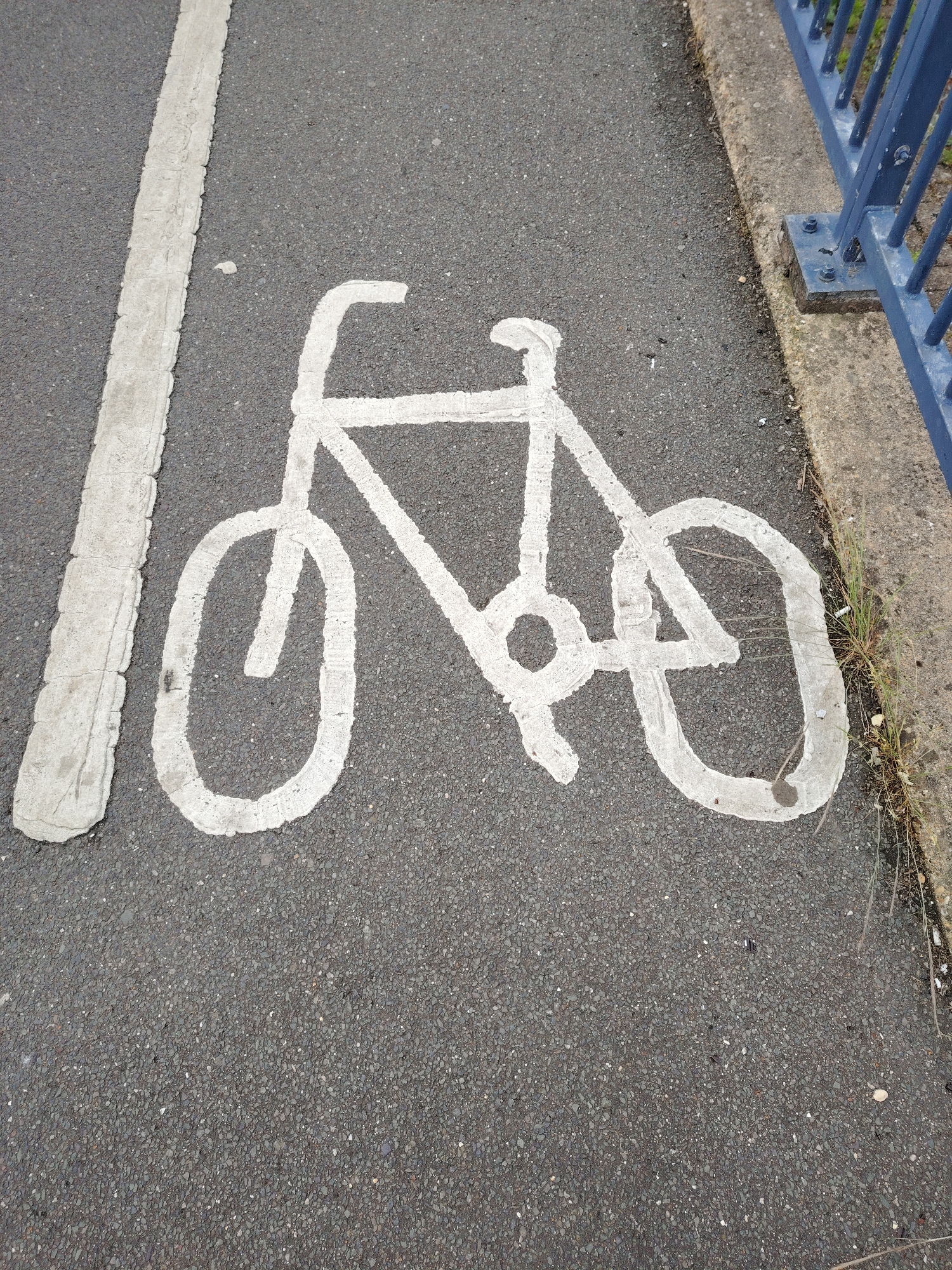 The image presents an overhead view of a dark grey asphalt surface, characteristic of a pathway or road. A thick, textured white painted line runs along the left side, exhibiting wear and minor cracking. Centered within the frame is a prominent white bicycle symbol, manually painted with some irregularities in its lines and displaying signs of weathering and dirt accumulation. To the right, a light grey concrete curb delineates the edge of the asphalt. Beyond the curb, a blue metal fence or railing is visible, anchored to the concrete base with bolts. Sparse, dry grass and small debris are present along the interface between the asphalt and the curb. No individuals or active subjects are present in the scene. The precise location within Peterborough, United Kingdom, cannot be determined from the visual content provided.