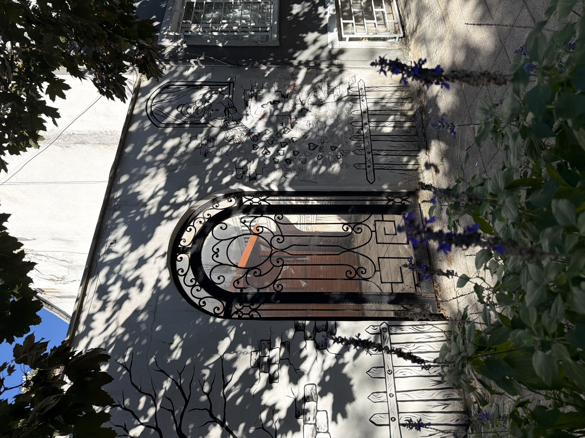 The image depicts a sun-drenched wall adorned with black and white drawings and graffiti. An ornate black metal gate stands in the center, allowing a view into a courtyard with a red-brick building. The wall features a mural of a stylized tree with branches casting shadows, a drawn picket fence, and various sketches including a cartoon of an elderly woman, hearts, and scribbled names. The building itself is light-colored with some visible cracks and weathering. To the right of the gate, a window with a metal grate is visible, and below it, another small window with a similar grate. In the foreground, out-of-focus purple flowers and green leaves add a touch of nature to the scene. The ground is paved with square tiles. The overall impression is an urban outdoor setting with artistic embellishments. The specific location is in the Moda neighborhood of Kadıköy, Istanbul, Turkey.