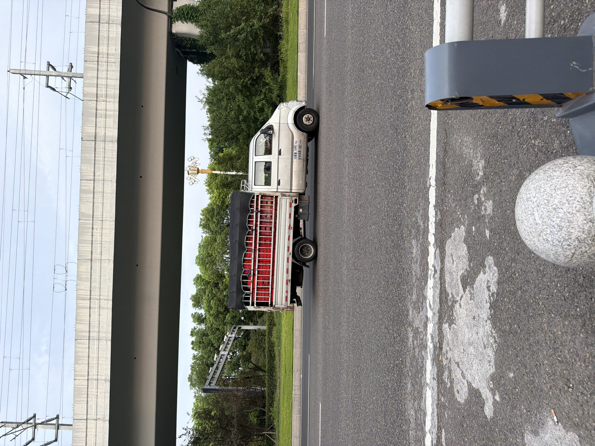 The image shows a light-duty truck driving on an asphalt road. The truck is silver and has a black tarp covering the bed, which is filled with red containers. The truck has Chinese text on its side, indicating its weight capacity. In the background, there is an elevated concrete structure with overhead power lines, likely an overpass or bridge. Lush green trees and vegetation line the road and beyond the overpass. The sky is overcast. In the foreground, part of a grey and yellow traffic barrier and a rounded granite bollard are visible. The scene appears to be in an urban or suburban area of Jiangjin, China, given the infrastructure and vehicle markings. No people are clearly visible or engaged in any discernible actions.