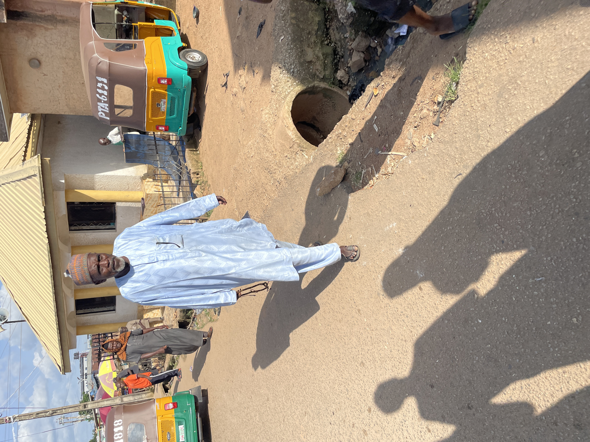 The image depicts a street scene in Gura Top, Nigeria, with a man in a light blue traditional outfit walking towards the camera. He has a beard and wears a patterned cap, and he carries a string of beads in his right hand. In the background, other people are visible, including a person in a grey garment walking away from the camera, and individuals near a colorful auto-rickshaw. The auto-rickshaw, with the registration number PTA-6131, is parked on the right side of the street. A building with a yellow roof is situated behind the main subject. The street is unpaved and dusty, with shadows cast by the people and a nearby structure. A large, concrete pipe, possibly a culvert, is present on the right side of the road, adjacent to a ditch containing debris and water. The environment suggests a typical urban or semi-urban setting in Nigeria.