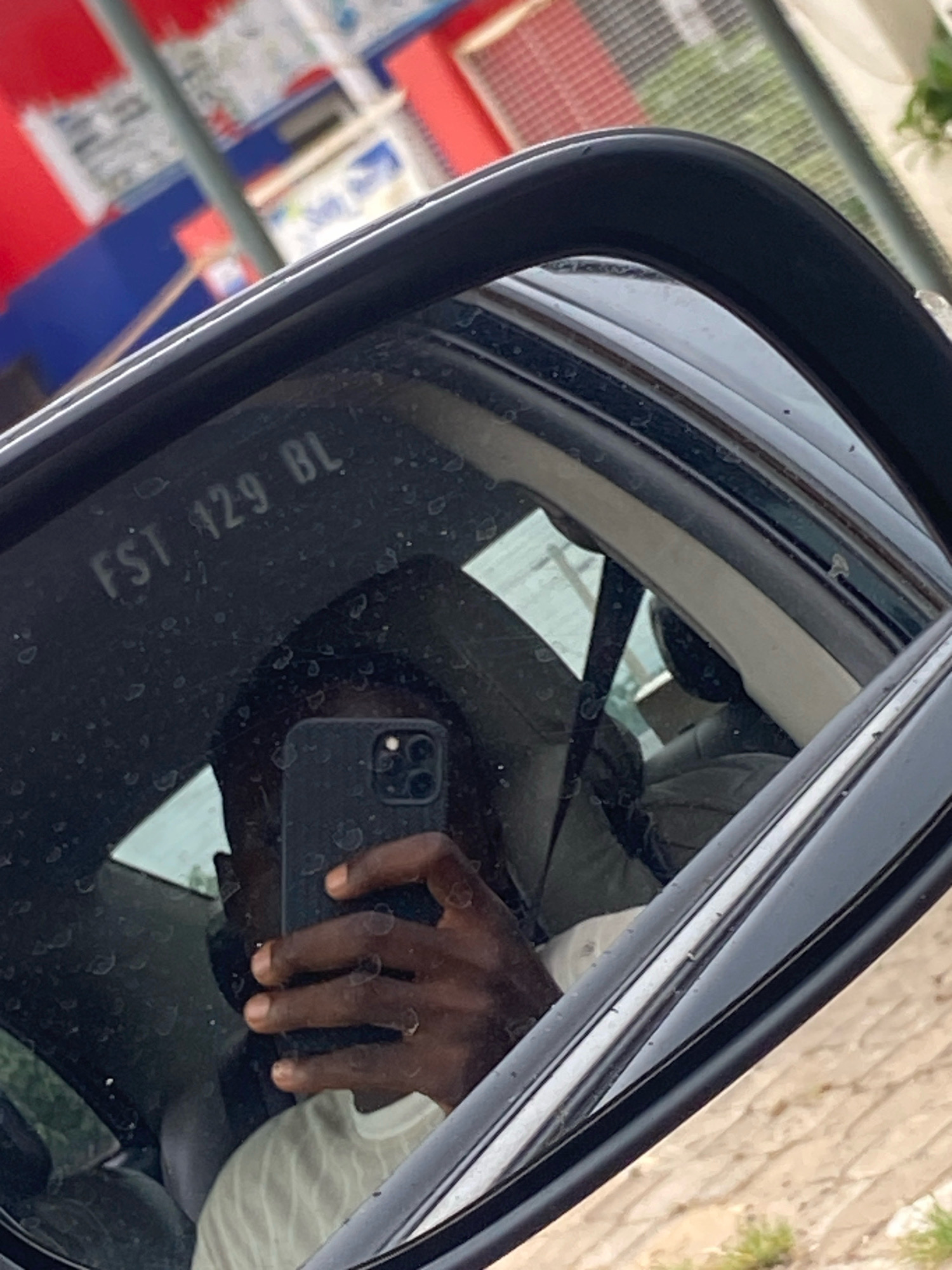 In the rearview mirror of a vehicle, a dark-skinned individual is reflected, holding a black smartphone to take a selfie. The individual is wearing a light-colored garment. The mirror itself is somewhat smudged. Reflected in the mirror, and visible in the background outside the vehicle, are elements of a street scene in Kaduna, Nigeria. These include buildings with red and blue facades, possibly shops or stalls, and a metal pole. The ground outside appears to be paved with soil and sparse vegetation.