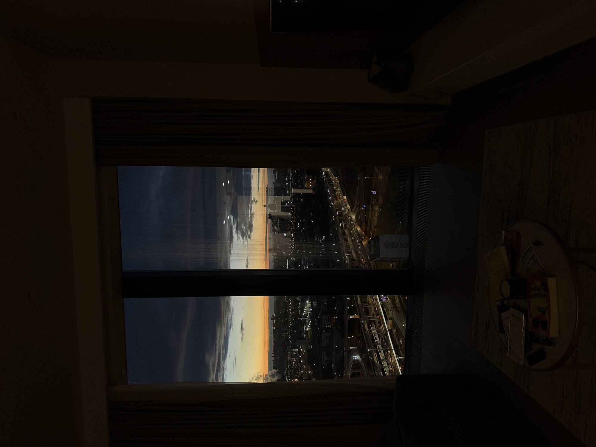 The image presents a night view from a window, likely an apartment or hotel room in Khimki, Russia. The window frames a cityscape at twilight, featuring artificial lights from buildings and street traffic. Visible are roads, possibly an expressway, with moving vehicle lights. Inside the room, the lighting is dim, with a round side table holding various objects, including books or magazines, a black mug or small container, and other miscellaneous items. The scene suggests a quiet, indoor setting overlooking an urban landscape at dusk.