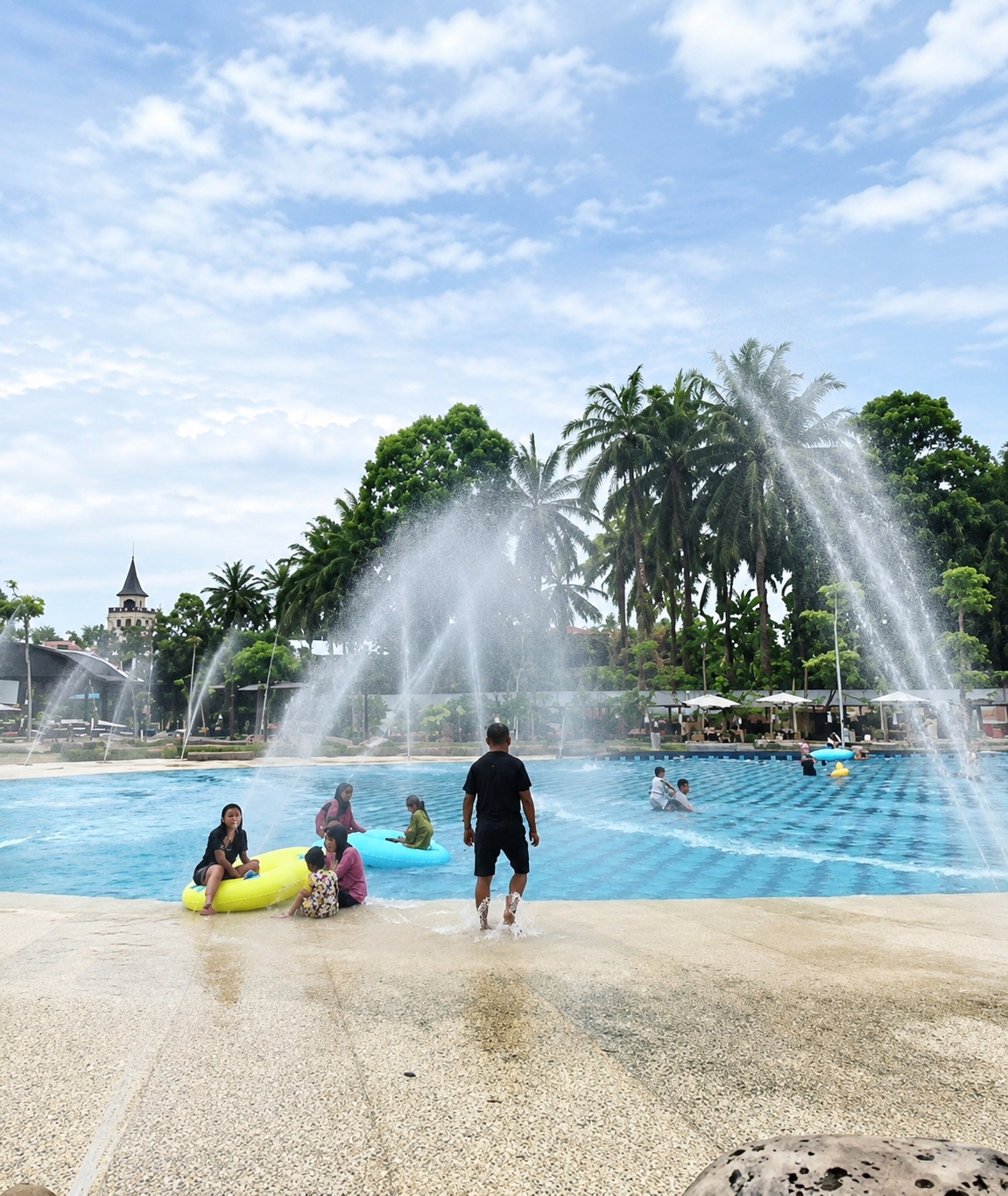 This image depicts a water park scene in what is identified as Eampat Kwalamencirim, Indonesia. The foreground shows a textured, light-colored surface with water splashing on it, indicating the edge of a pool. In the middle ground, a large blue swimming pool is visible, with multiple water jets creating spray and mist. Several people are in and around the pool.
In the immediate foreground of the pool's edge, a Black man wearing black shorts and a black t-shirt is walking away from the camera, with his feet visible in the splashing water. To his left, a group of people are near the edge of the pool. A woman in a black top and dark pants is sitting on a yellow inflatable ring. Beside her, a child is sitting on the ground. Further to the left, another woman in a pink top and dark pants is also sitting near the water. A young child is sitting on a green inflatable ring.
In the mid-ground of the pool, several individuals are swimming or floating. Two men are visible further back in the water. Another person is on a yellow inflatable ring.
The background features lush green trees and palm trees, characteristic of a tropical environment. A dark, tower-like structure with a pointed roof, resembling a castle or a distinctive building, is visible on the left side of the background, nestled amongst the trees. Umbrellas and seating areas are also visible in the distance near the pool. The sky above is bright blue with scattered white clouds. The overall impression is a lively recreational setting on a sunny day.
