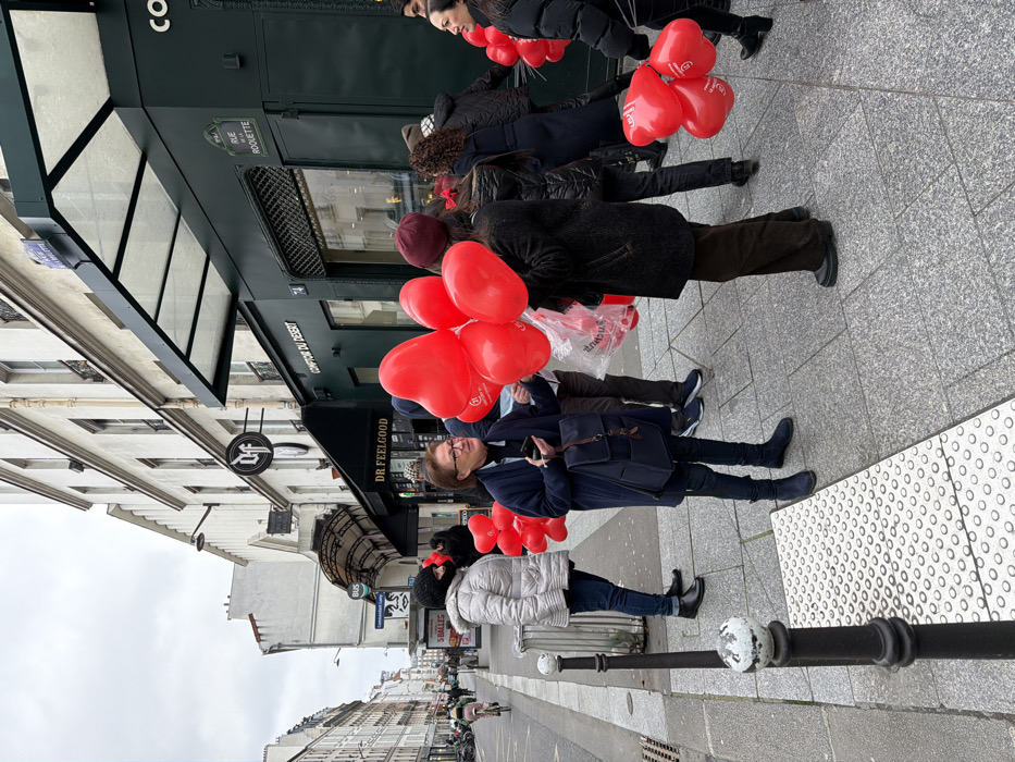 The image depicts an urban street scene on Rue de la Roquette in Paris, France, on an overcast day. The focal point is a gathering of individuals on a granite-paved sidewalk in front of commercial establishments.
A dark green storefront, labeled "COMPTOIR DU DESERT" at street number 74, features a dark awning and a visible "RUE LA ROQUETTE" sign. Adjacent to it, another shop with a lighter facade displays a circular "DF" logo and "DR FEELGOOD" signage.
Numerous red heart-shaped balloons are present, held by multiple people or contained in a clear plastic bag. A woman in a dark blue coat and glasses, holding a smartphone and a dark blue backpack, stands centrally, looking towards her right. To her right, a person in a dark coat and maroon hat holds a large cluster of these balloons, some marked with a white logo. Another woman in a silver puffer jacket and black hat stands to the left, also looking right.
Several other individuals are visible, mostly dressed in winter attire, clustered around the shop entrance, many also holding red balloons. In the background, the street recedes with multi-story Parisian buildings. A cyclist is visible further down the street, and various street furniture including black bollards, a tactile paving strip, and a bus stop sign are present on the sidewalk.