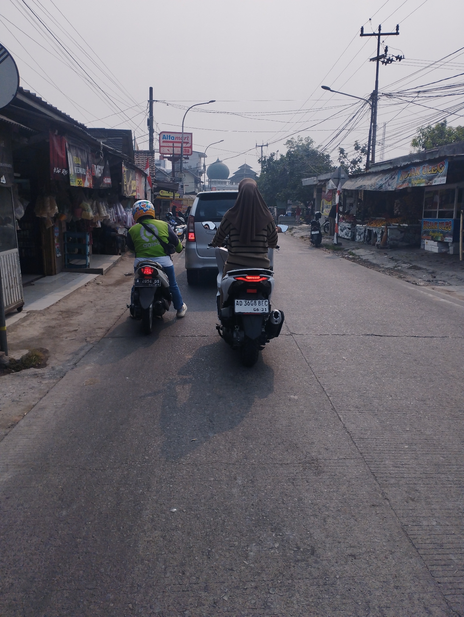 The image depicts a street scene in Kramatwatu, Indonesia, with two individuals riding motorbikes. The person in the foreground on the right is riding a dark-colored motorbike with a license plate reading "AD 3608 BEC". They are wearing a light brown headscarf and a striped long-sleeved shirt. The person on the left is on a lighter-colored motorbike with a license plate reading "A 2056 DS". They are wearing a green jacket with a yellow logo and a helmet with a cartoon character design. Both individuals are moving away from the camera down a paved road. The road is lined with shops and stalls on both sides. On the left, there are shops with awnings and various items displayed outside. A sign for "AlfMart" is visible in the distance. On the right, more shops with awnings are present, and in the background, a green dome structure, possibly a mosque, can be seen. Overhead, a complex network of power lines stretches across the sky. The sky is overcast, suggesting either early morning or late afternoon.