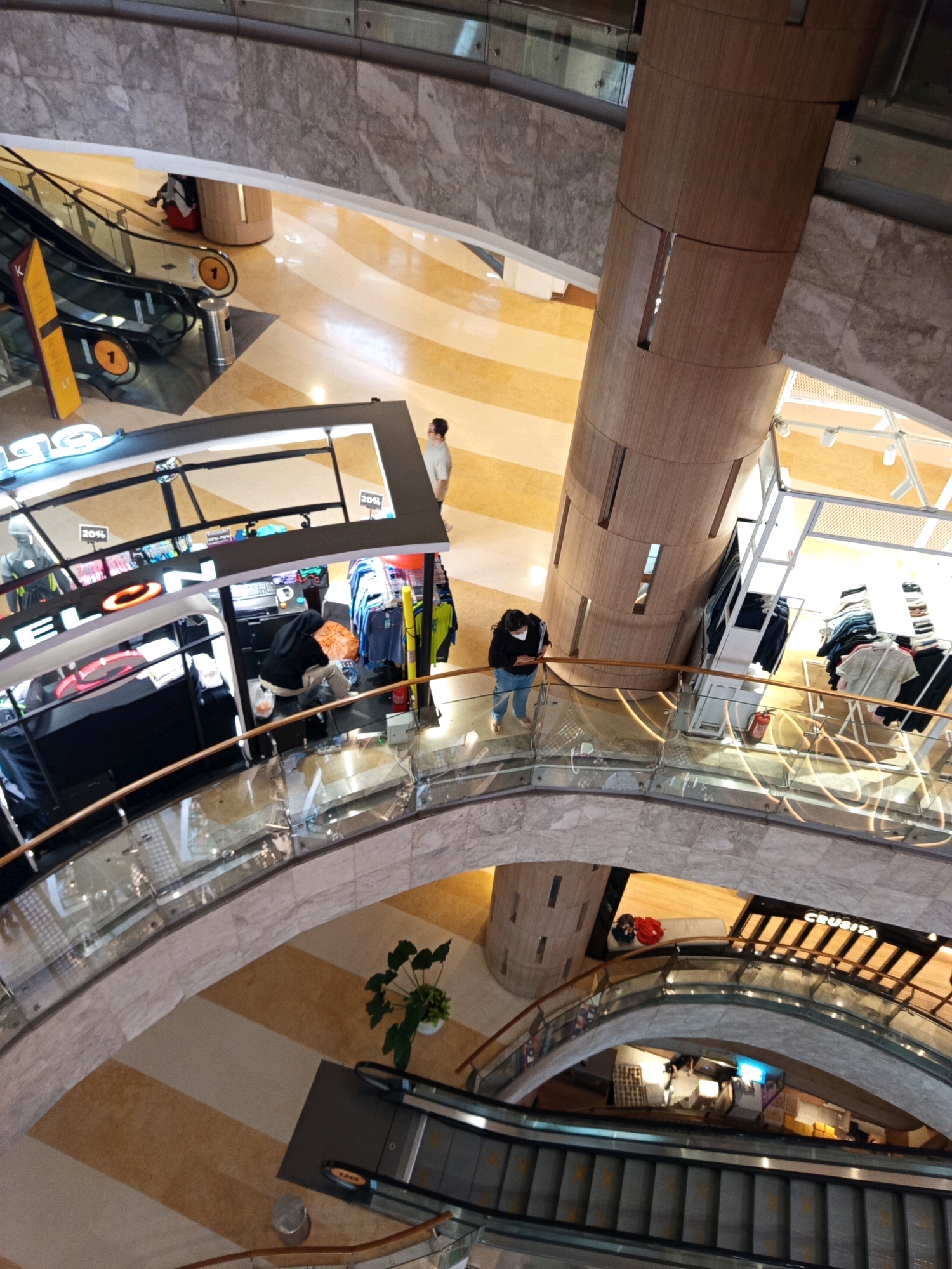The image depicts an interior view of a multi-level shopping mall in South Jakarta, Indonesia, likely taken from an upper floor looking down. In the foreground, a curved glass and marble railing lines a walkway. Below this, escalators are visible, with the lower ones appearing to be in use. A person is seen on one of the escalators.
Further into the mall, a central, cylindrical wooden pillar dominates the space. To the left of the pillar, a store with a black facade and neon signage is partially visible, with mannequins and clothing racks displayed. A person is seen tending to a display inside this store.
To the right of the wooden pillar, another store is visible, featuring racks of clothing. A fire extinguisher is mounted on a wall in this area. On the walkway above the escalators and next to the wooden pillar, two individuals are walking.
The mall's flooring features a patterned design of beige and light brown stripes. Lighting fixtures are visible on the ceiling. The overall impression is of a modern, well-lit retail environment.