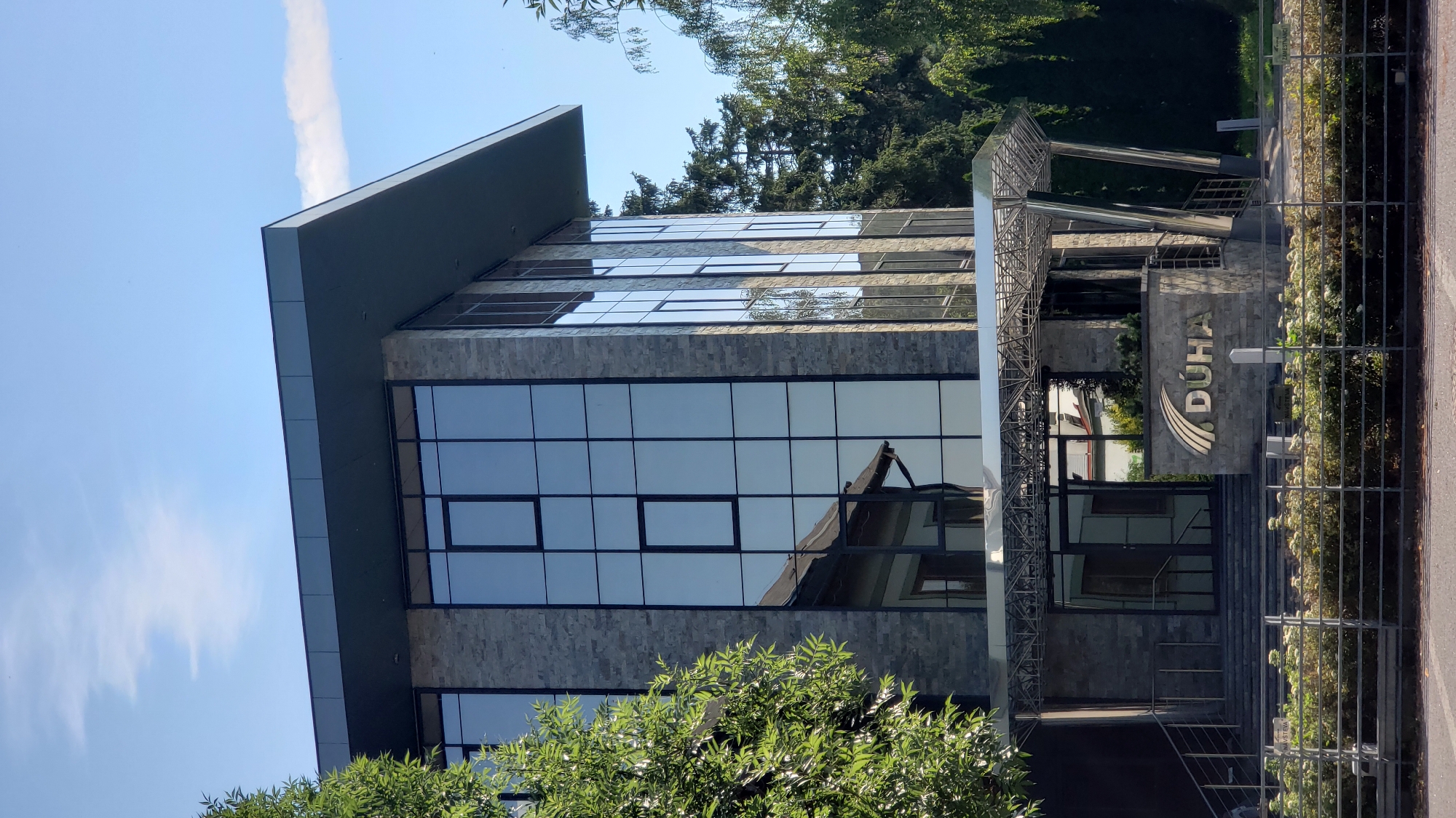 The image shows a modern office building with a glass facade and stone accents. A metallic awning extends over the entrance, which is flanked by stairs and a stone wall bearing the logo "DÚHA". The building is surrounded by greenery, including trees and bushes. The sky is blue with a few wispy clouds. The scene appears to be a daytime shot, with clear weather. The building is identified as the DÚHA office building located in Prešov, Slovakia.
