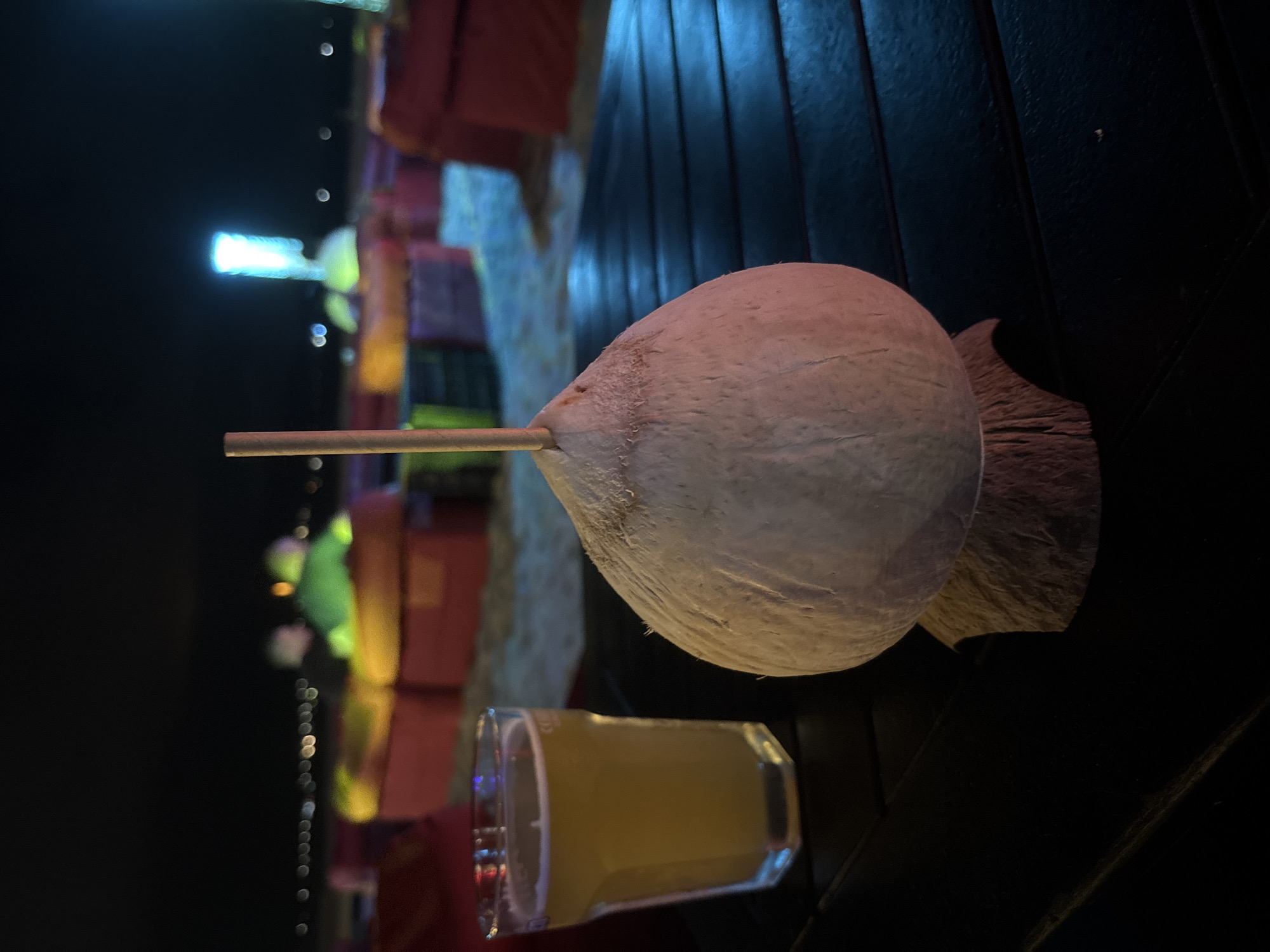 At night in Đà Nẵng, Vietnam, a young coconut drink with a straw and a shot glass of amber liquid are on a dark, horizontal wooden table. Beyond these items are out-of-focus, colorful decorations and a line of lights, suggesting a festive or celebratory atmosphere.