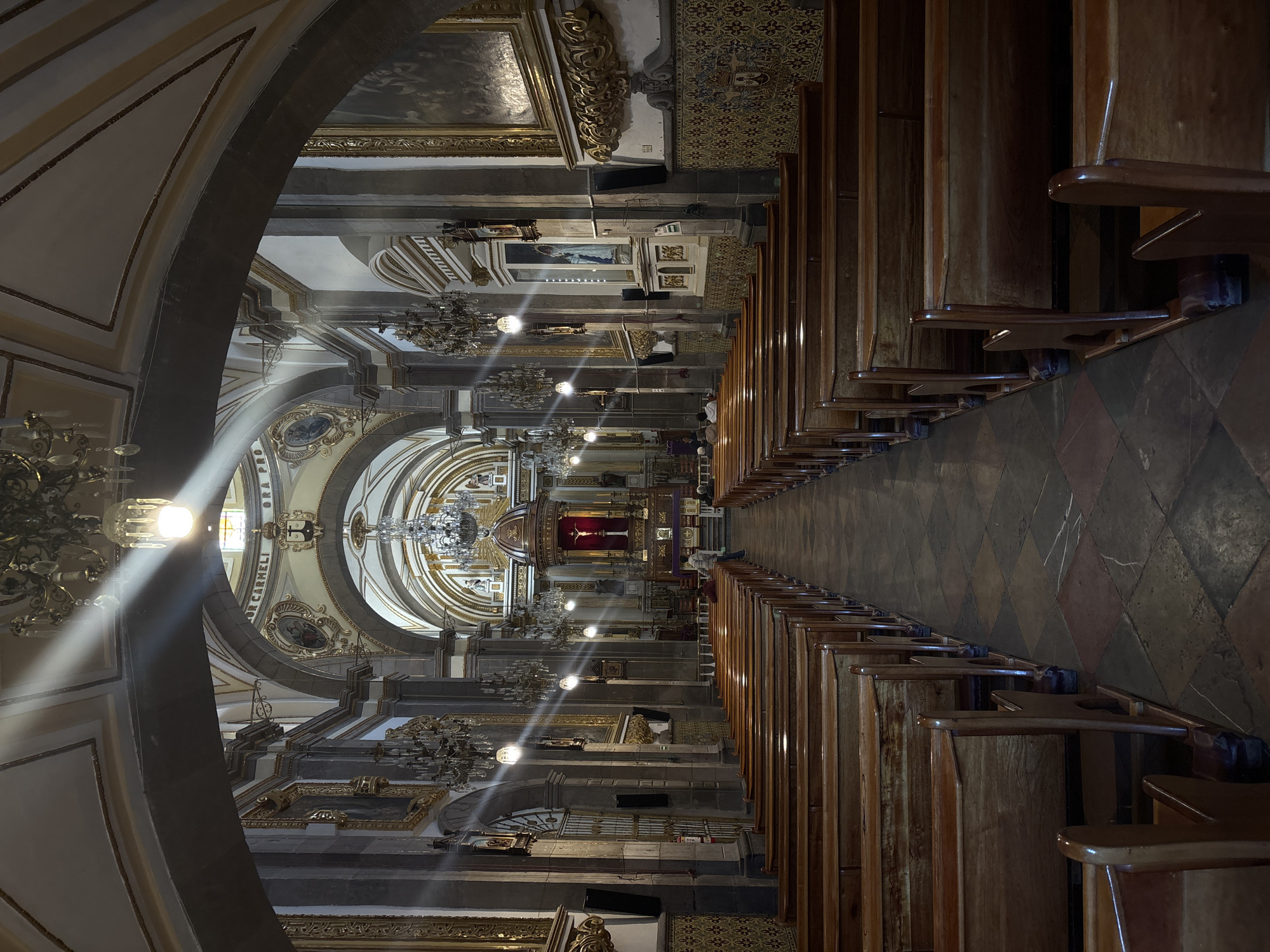 This image shows the interior of a church, with rows of wooden pews leading toward the altar. Chandeliers hang from the ceiling along the nave. The altar area features ornate gold trim and a crucifix. Two people are seen walking towards the altar and a women is kneeling in prayer. The image is taken from within an arched entryway. The location is likely in the city of Puebla, Mexico, based on the architectural style and decorative elements. The floor is tiled with a diamond pattern.
