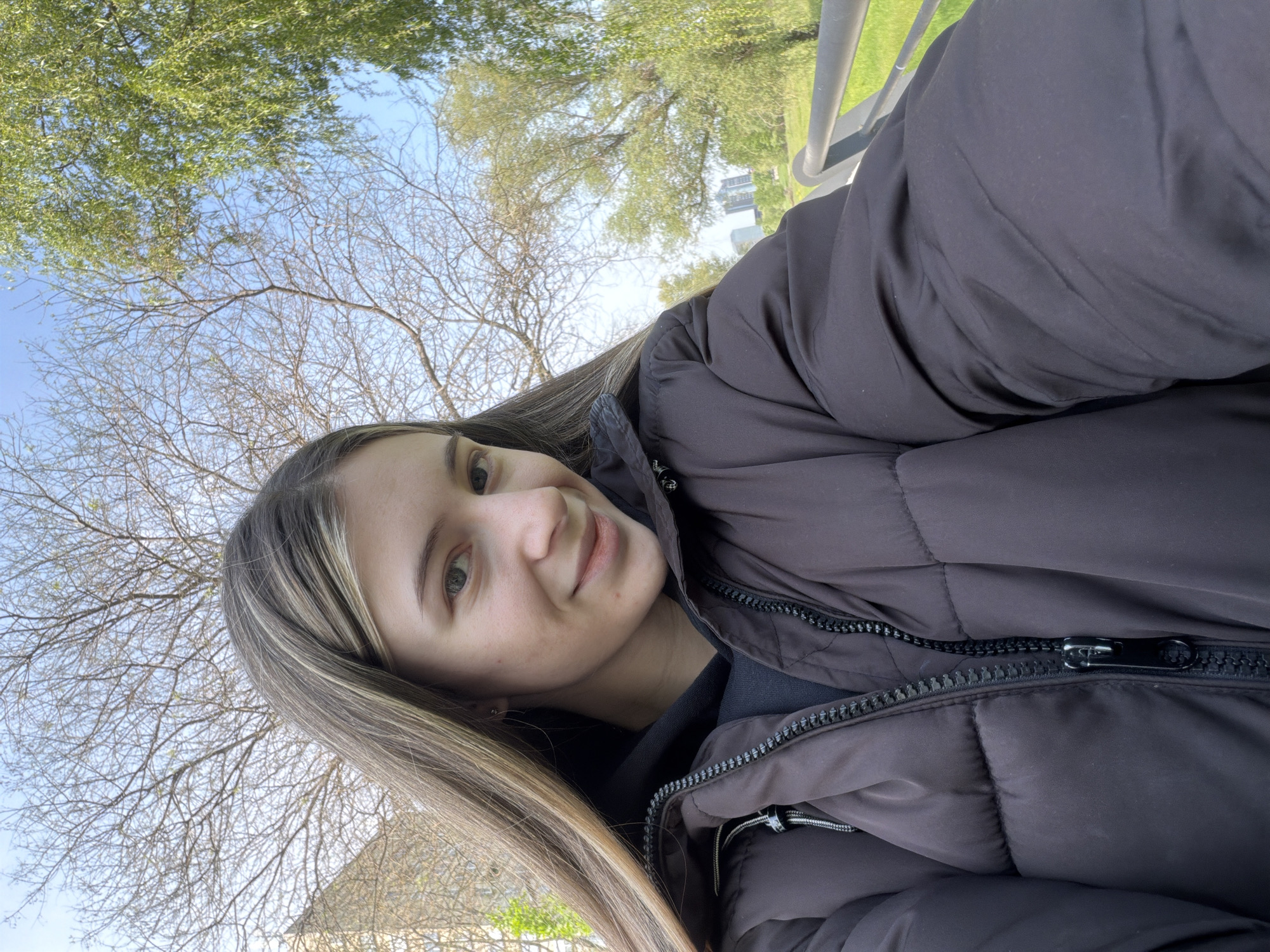 The image, rotated sideways, features a young woman taking a selfie outdoors in what appears to be a park or green urban area in Plovdiv, Bulgaria. The setting suggests daytime under a clear blue sky.
The woman, likely in her late teens or early twenties, is positioned in the center-right of the frame, looking towards the camera with a soft, pleasant smile. She has long, straight hair with light brown and lighter, possibly highlighted, streaks, and light-colored eyes. She is wearing a dark, likely charcoal grey or brown, puffer jacket with a visible black zipper and a metallic pull tab. A dark-colored top is visible underneath her jacket.
The background consists mainly of trees and sky. Above and to the left of the woman, some trees are full of vibrant green leaves, while others towards the bottom-left are bare, showcasing their intricate branch patterns against the bright blue sky. A light-colored metal railing is visible on the far right. In the distance, beyond the trees and grass, faint outlines of urban buildings can be discerned. The bright, even lighting suggests it is daytime. No visible text is present in the image.