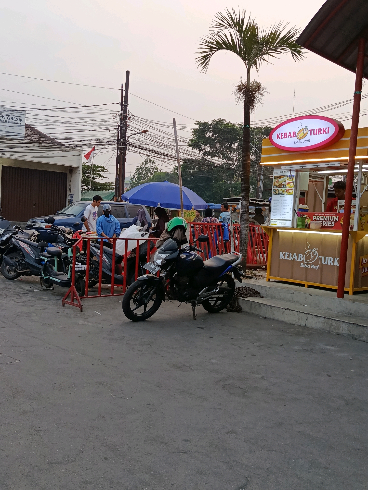 The image depicts an outdoor scene at dusk, likely in Bekasi, Indonesia. In the foreground, two motorcycles are parked on an asphalt surface. A red barrier is positioned between the motorcycles and a food stall. The food stall, branded with "KEBAB TURKI Baba Rafi," features bright yellow and orange signage and an illuminated menu. A young man in a red shirt is visible behind the counter, possibly serving customers. In the middle ground, several people are gathered around another stall with a blue umbrella. A dark blue SUV is parked behind them, and another motorcycle is partially visible to the left. Power lines crisscross the sky, and a palm tree stands tall next to the kebab stall. A building with a sign reading "GRES... IA TI..." is visible to the left. The overall atmosphere suggests a bustling commercial area.