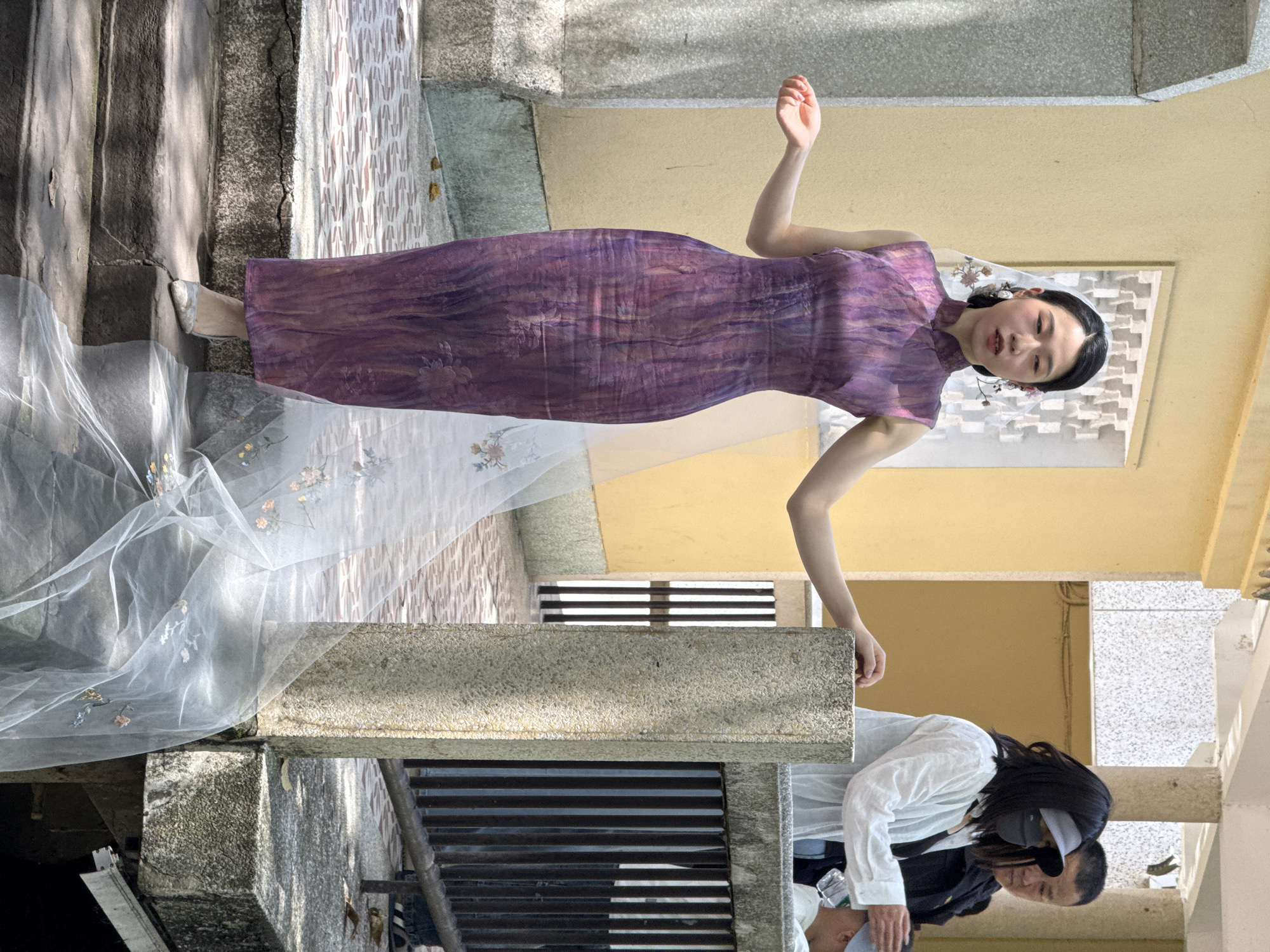 A woman in a purple dress and white veil poses in what appears to be the Yuzhong District of China. The woman is positioned on the left side of the image, her body oriented diagonally. The background features architectural elements, including steps and walls. Another person wearing a white shirt is present in the bottom right corner of the image.