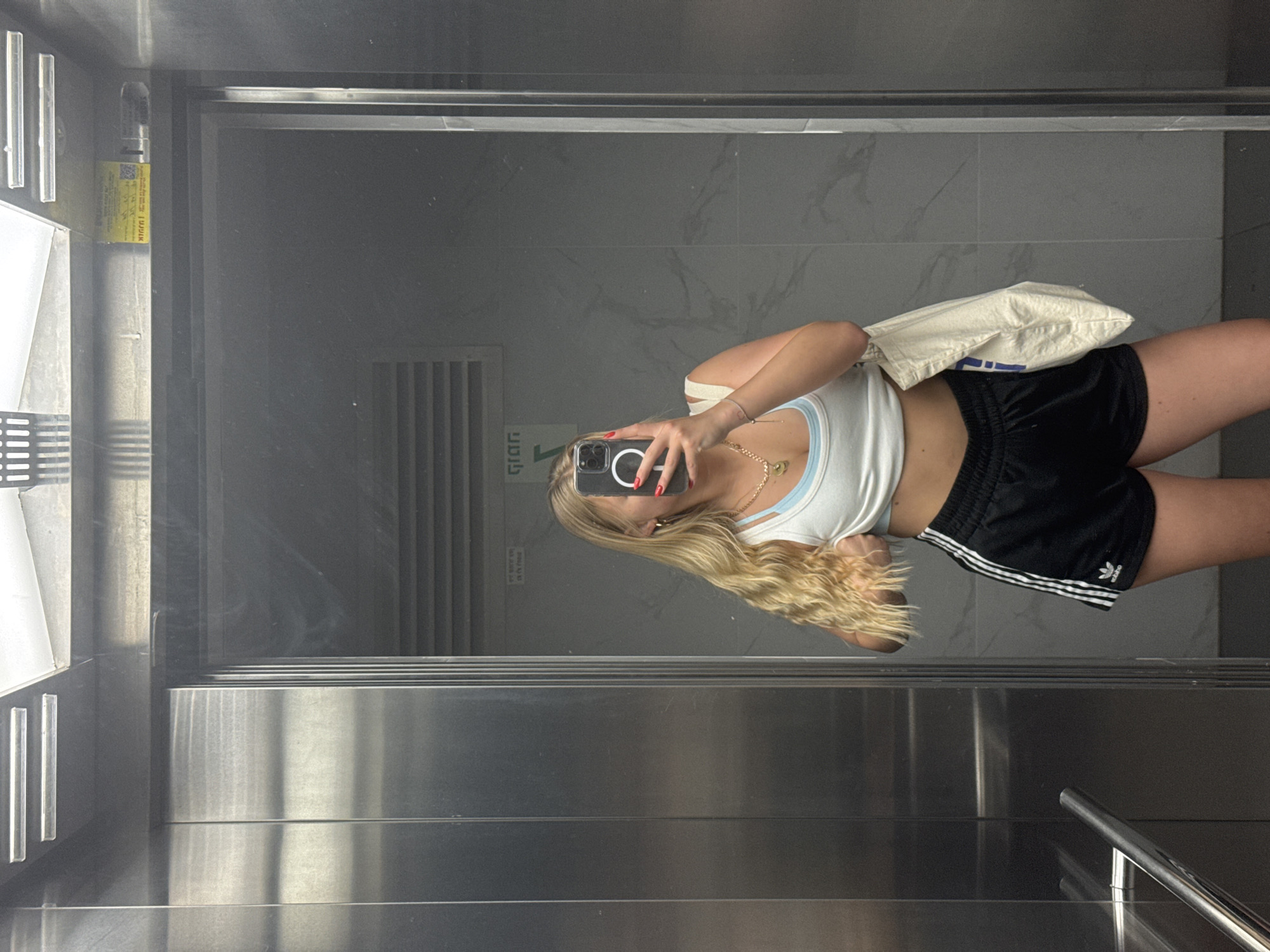 A woman is taking a selfie in an elevator. She is wearing a white tank top, black Adidas shorts, and gold jewelry. She has long blonde hair. The elevator has metal walls and a marble-like back wall. The location is in the city of Unknown, Unknown.