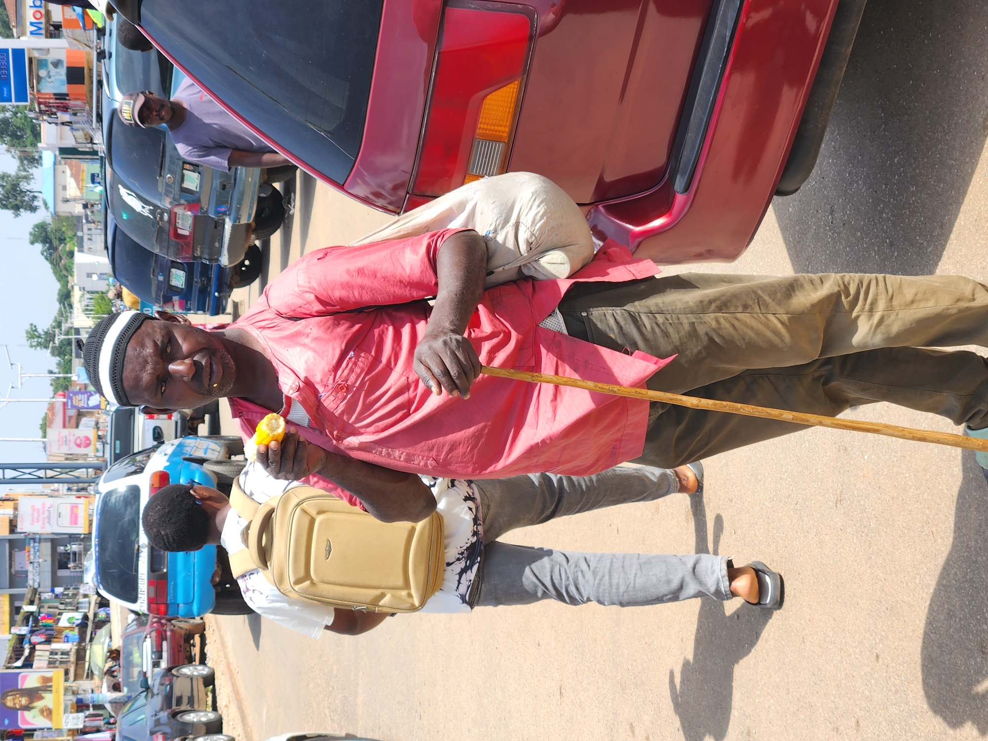 In an outdoor, urban environment, a man wearing a red shirt, brown pants, and a white and black cap stands in the foreground, holding a wooden stick and eating corn on the cob. He has a light-colored bag slung over his left shoulder. To his left, a person in a white shirt and gray pants, carrying a light-brown backpack, walks away from the camera. Behind them, several vehicles are parked on the asphalt road, including a blue and white van and a dark-colored SUV. In the background, buildings with various signs are visible, suggesting a commercial area. The lighting indicates it is daytime, with shadows cast on the ground.