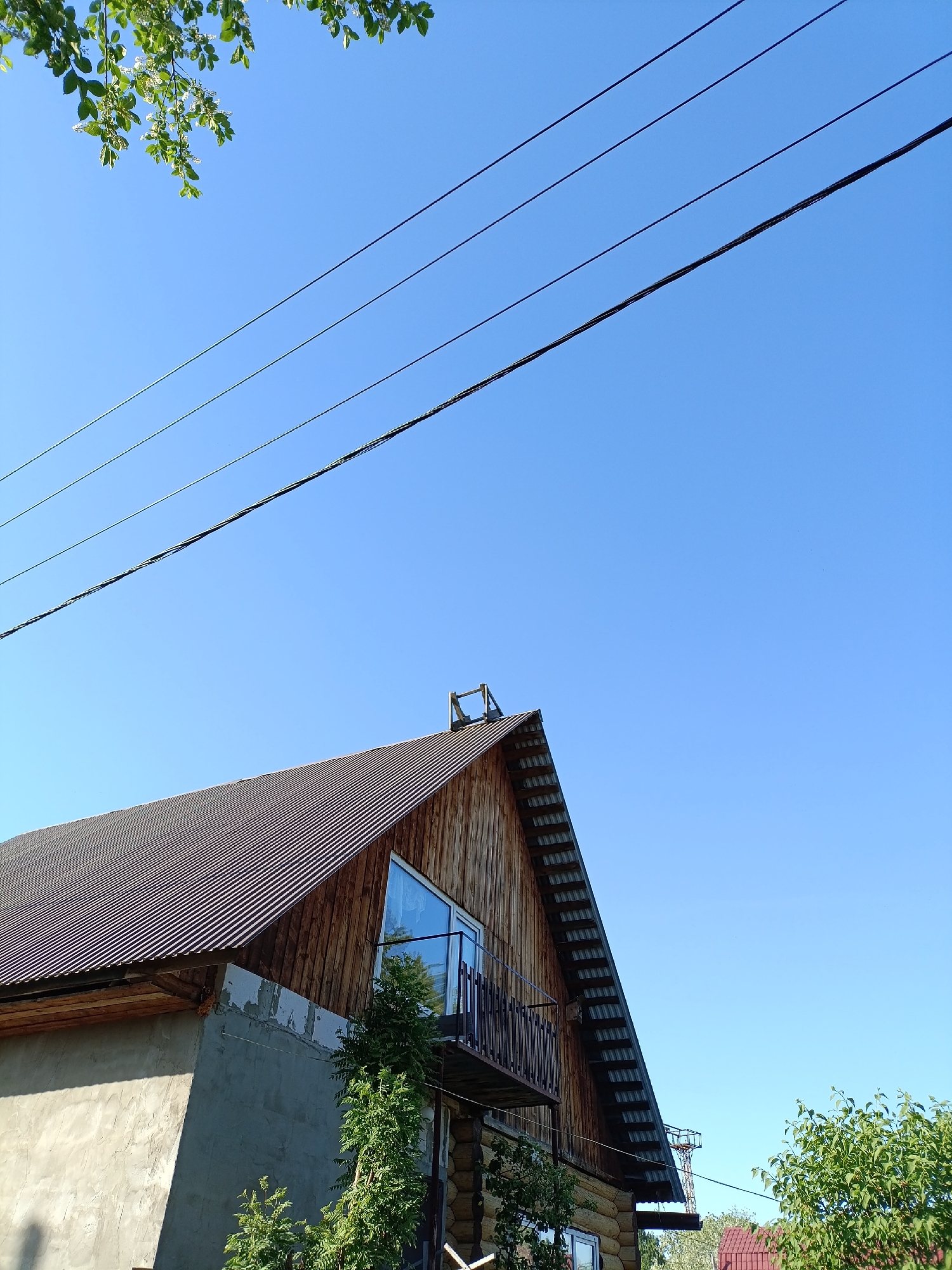 The image presents a view of a building, likely a residential structure, situated in what appears to be a suburban or rural setting. The building features a steeply pitched roof covered with corrugated metal. A prominent gable end is visible, with wooden siding and a triangular window. A small balcony is attached to the upper level. The exterior of the structure combines modern materials like the metal roof and triangular window with log construction. The wall section nearest to the camera looks to be concrete or stone.
The sky above the structure is clear and blue, with power lines bisecting the view. Green foliage is partially visible in the corners of the image, implying the presence of trees or shrubbery.
While the location in Chaikovsky, Russia is mentioned, no specific architectural characteristics that uniquely link this building to that location are evident from the image. The style blends modern design elements with vernacular log construction, a common style in regions with access to timber.