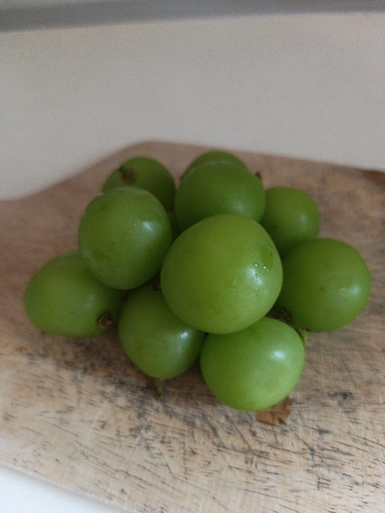 A pile of green grapes rests on a wooden cutting board. The grapes are round and plump, with a smooth, slightly waxy surface. Some of the grapes have small stems attached. The wooden cutting board has a weathered appearance with visible grain lines and some dark marks. The background is out of focus and appears to be a light-colored wall or surface. The scene is lit from above, casting subtle shadows on the grapes and the board. There are no people or any other objects visible in the image. This image was taken in Karangploso, Indonesia.