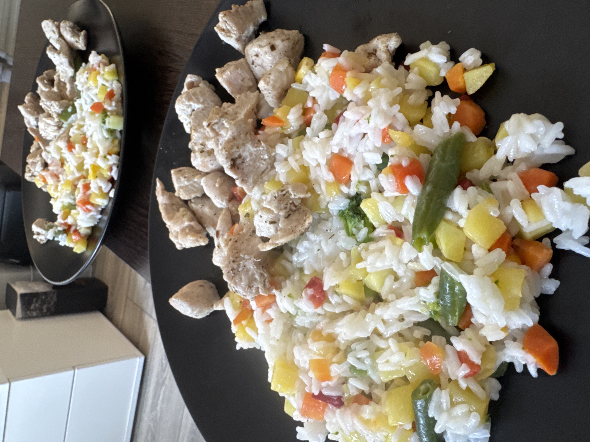 Two black plates of food are on a dark brown surface. Each plate contains cooked rice mixed with diced vegetables, including carrots, yellow squash, tomatoes, and green beans. Diced pieces of cooked chicken are arranged alongside the rice and vegetables. The plates appear to be in a residential setting near a white cabinet. It is not possible to determine if this is Murino, Russia from the image.