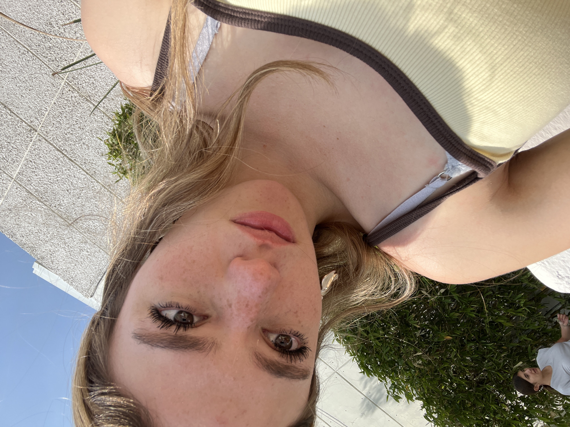 A young woman with light brown hair and prominent freckles captures a close-up selfie outdoors on a bright, sunny day. She is looking directly into the camera, with her lips slightly parted. She wears mascara on her eyelashes, and a white earbud is visible in her right ear. Her attire consists of a light yellow ribbed tank top with a dark brown trim.
The background reveals a clear blue sky on one side and vibrant green foliage, likely from trees or bushes, on the other, suggesting an open-air setting. A light-colored, textured surface, possibly pavement or tiles, is also visible in the background. Strong sunlight illuminates the scene, indicating daytime, likely mid-day or afternoon. A second person with dark hair, wearing a light-colored t-shirt, is partially visible in the bottom right corner of the frame, facing away from the camera. The woman appears calm and relaxed while taking the picture. The location context suggests this scene is in Puebla, Mexico.