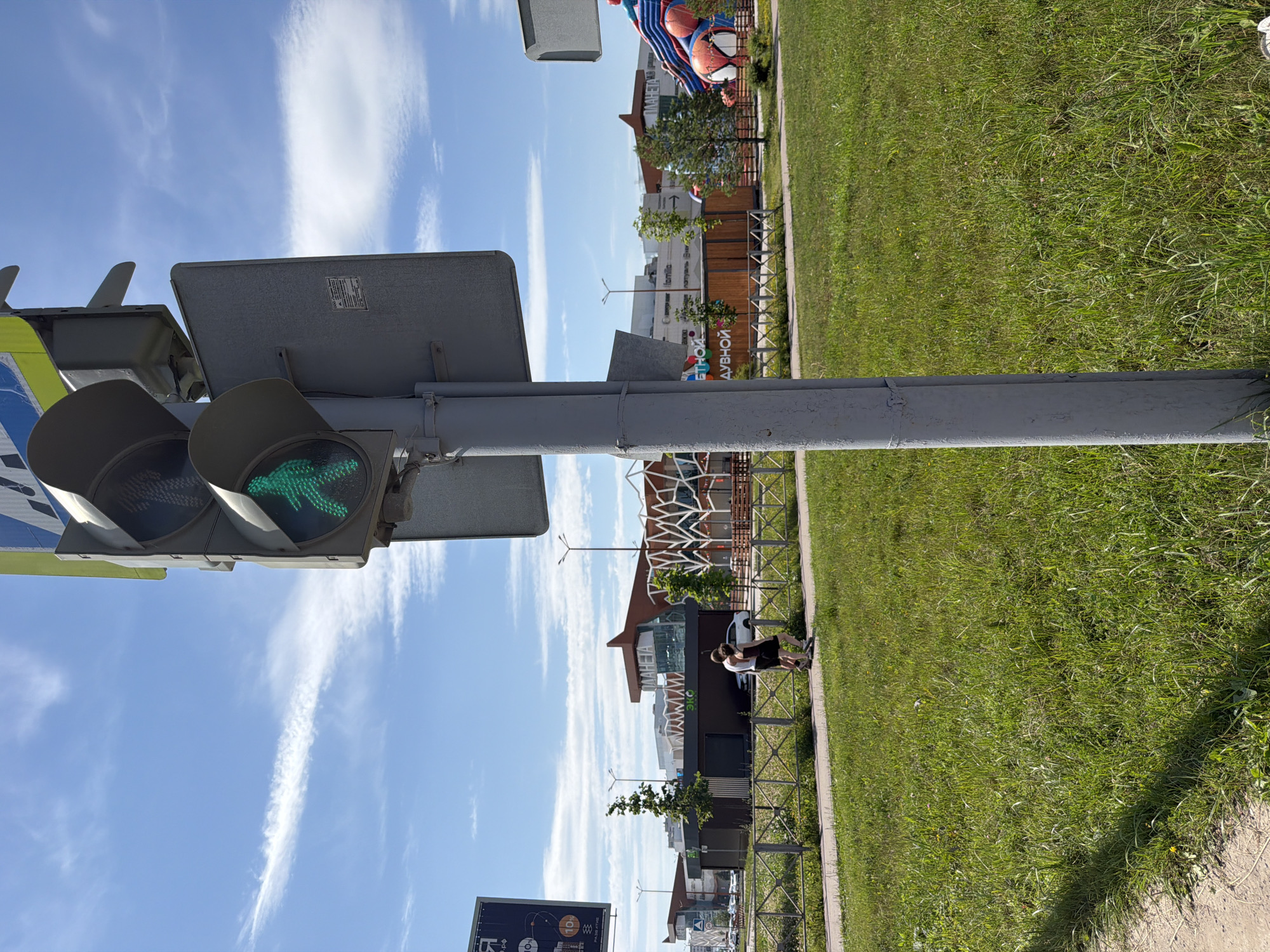A green pedestrian signal is illuminated, indicating it is safe for pedestrians to cross. In the background, two individuals, a man and a woman, are walking on a sidewalk beside a grassy area. Beyond the sidewalk, a fence separates the green space from a road or pathway. In the distance, several modern buildings with unique architectural designs are visible, including one with a prominent sign that reads "ПЛАНЕТА" (Planeta). A large inflatable Spider-Man attraction is also present near the buildings. The sky above is blue with scattered white clouds. The scene appears to be in a commercial or recreational area, possibly a shopping center or park, in the city of Perm, Russia.