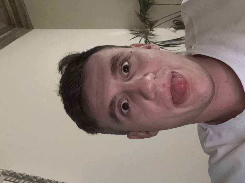 A male subject is depicted in a close-up, head-and-shoulders indoor view. He has short, dark hair, wide-open eyes, and his tongue is extended from his open mouth. He is wearing a white t-shirt. The background consists of a plain, light-colored wall. A decorative architectural molding is visible in the upper right quadrant, and foliage from a green potted plant is present on the right side. The scene is situated in Venice, Italy.