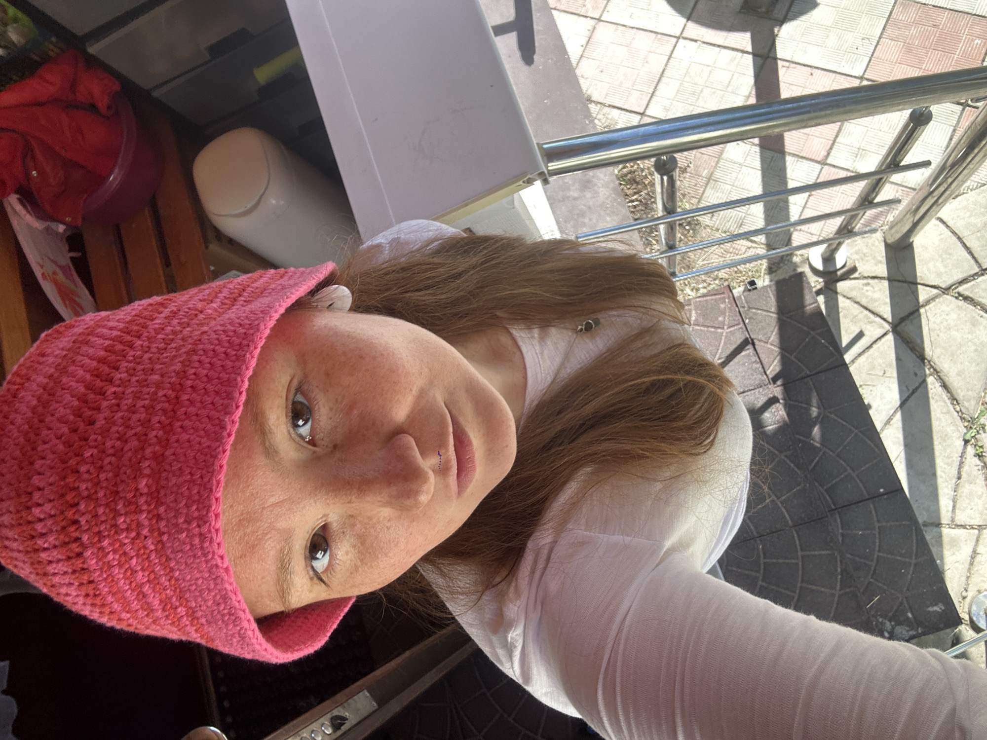 This media file is a high-angle selfie-style portrait of a person, rotated approximately 90 degrees clockwise, taken outdoors in Nizhny Novgorod, Russia.
The main subject is a person with light brown or auburn hair and visible freckles on their face. They are looking directly at the camera with a neutral, calm expression, and have dark eyes. The person is wearing a knitted pink hat or wide headband and a white long-sleeved top. A subtle lip piercing is visible below their nose, and a small earring or piercing can be seen in their right earlobe.
The background is split between a cluttered indoor or semi-outdoor space on the left (bottom of the original frame) and an outdoor setting on the right (top of the original frame). On the left, there's a wooden structure (possibly furniture), a red folded garment, a purple bowl, a white cylindrical container, and partially visible gray drawers or shelves. On the right, bright sunlight illuminates a paved area with square and circular tile patterns, adjacent to silver metal railings. This suggests the photo was taken on a balcony, porch, or elevated outdoor area.
The strong, distinct shadows cast on the tiled ground indicate bright, sunny weather, likely during midday or early afternoon. There is no visible text in the image.