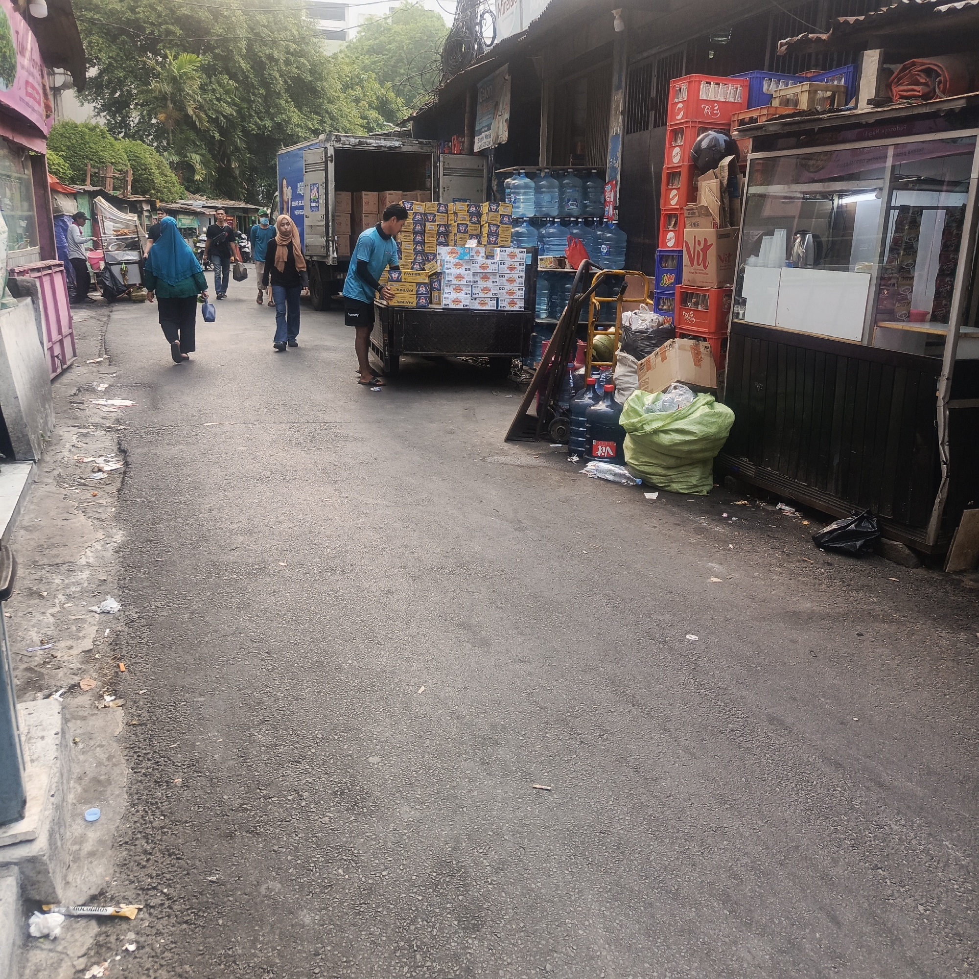The image depicts a street scene in South Jakarta, Indonesia, characterized by a narrow asphalt road lined with various roadside establishments and vendors. In the foreground, the road is somewhat worn and shows signs of litter. Several people are walking along the road. On the left side of the street, there are low-rise structures, some with pink accents, appearing to be shops or stalls. Further down the street, a large delivery truck is parked with its back open, revealing stacks of cardboard boxes. A man in a blue t-shirt is actively unloading these boxes. Adjacent to the truck, there are numerous large blue water dispenser jugs, some stacked on shelves.
To the right of the road, there are more stalls and displays. Prominently visible are stacks of red and blue crates, some filled with bottles. A glass-fronted kiosk or shop is also present, with what appears to be food or drinks being displayed inside. A large green bag, likely containing trash or goods, is placed on the ground near the water jugs. A black trash bag is also visible on the right side of the road. The overall impression is of a busy, functional commercial area, likely a market or a street with numerous small businesses, typical of some areas in urban South Jakarta. Trees and foliage are visible in the background, suggesting a tropical climate. The lighting indicates it is daytime, possibly overcast or early morning due to the diffuse light.