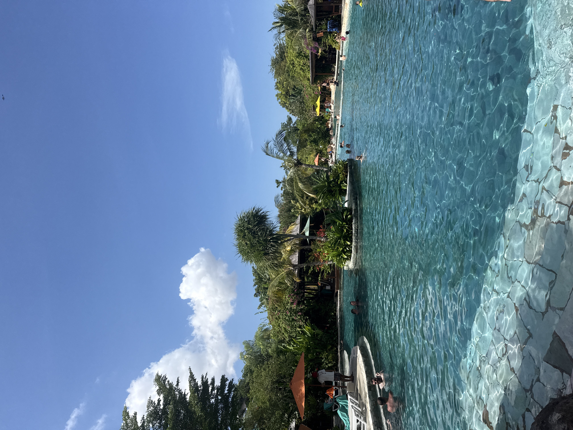 The image displays a vibrant outdoor scene at a tropical resort or recreational area, likely in Kuta, Indonesia, under a clear midday sky. The composition is primarily divided between a bright blue sky on the left and a large, inviting swimming pool with lush tropical vegetation on the right.
The central subject is a spacious swimming pool filled with clear, turquoise water, exhibiting shimmering reflections of sunlight on its surface. The pool's edge and steps are crafted from irregular, light-colored flagstones or tiles, visible beneath the water. Several individuals are present in the pool, engaged in leisure activities such as swimming, wading, or simply relaxing in the cool water. Their forms are partially obscured by the rippling surface.
Surrounding the pool is dense, verdant tropical foliage, including various palm trees, tall leafy trees, and numerous bushes, creating a lush, secluded environment. Small structures with dark roofs, possibly bungalows or service buildings, are nestled within this greenery, adding to the resort aesthetic. Colorful sun umbrellas, including a prominent yellow one and an orange one, are dotted along the poolside, providing shade. White lounge chairs are also visible on the pool deck.
The weather is distinctly sunny, with a brilliant blue sky punctuated by a few wispy white clouds, suggesting optimal conditions for outdoor activities. The strong sunlight casts crisp shadows and highlights the vivid colors of the scene. The overall mood conveyed is one of relaxation, vacation, and enjoyment in a beautiful tropical setting. No discernible text is visible in the media file.