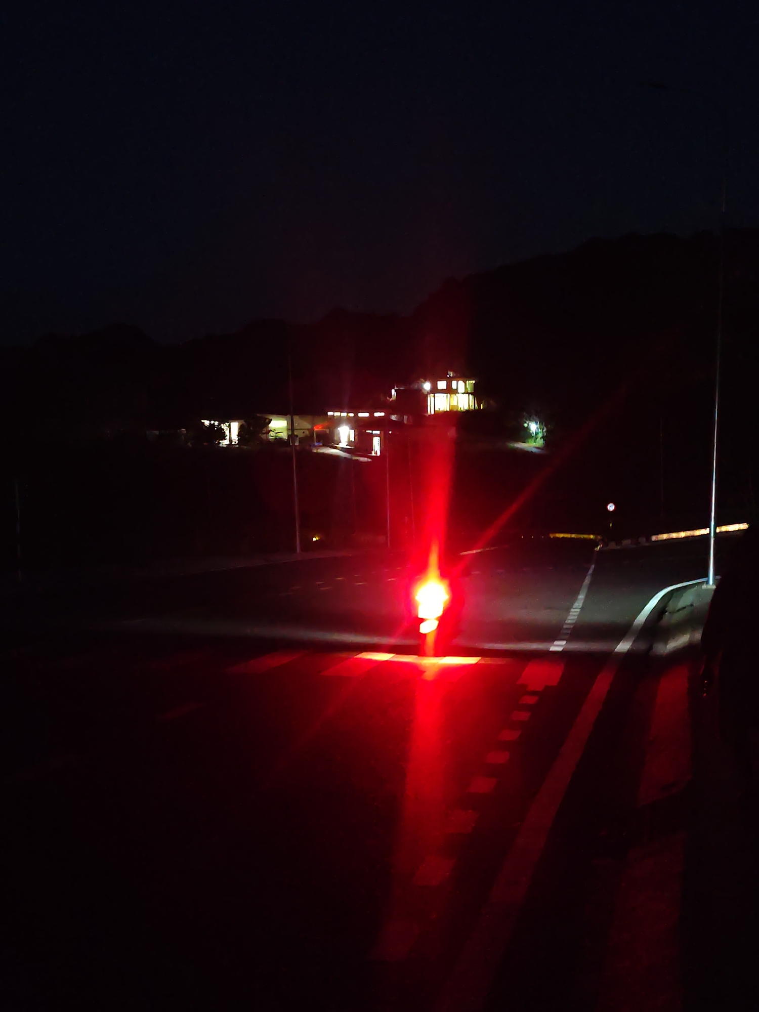 The image depicts a dark, rural scene at night, with a building complex illuminated in the distance against a backdrop of hills. The foreground shows a road with white dashed lines. A bright red light, likely from a vehicle, is prominent in the lower center of the image, casting a strong glow and reflections on the road surface. There are no discernible people or specific landmarks that would indicate Da Lat, Vietnam. The overall atmosphere is dimly lit and quiet.