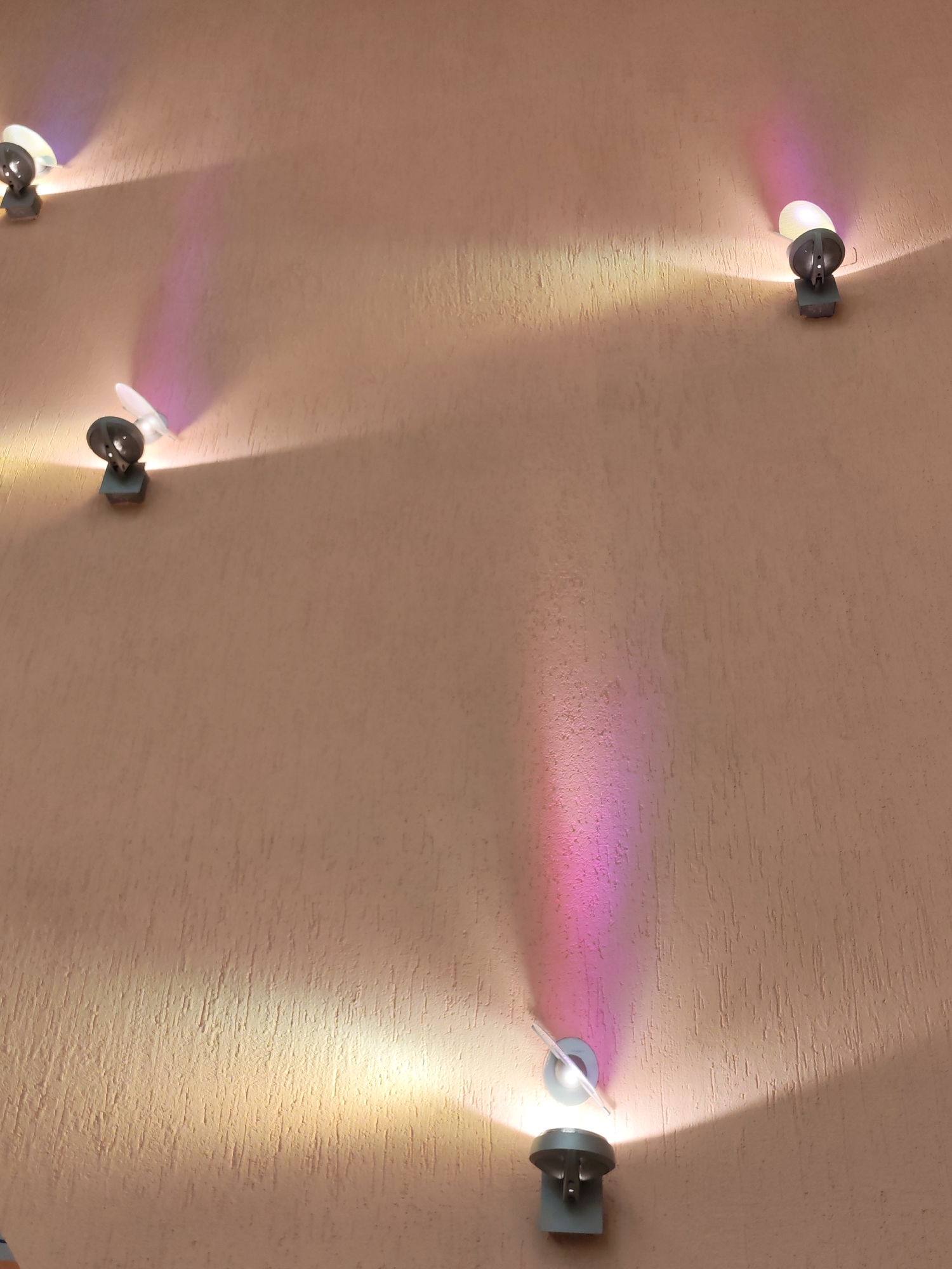 The image features a warm-toned, textured wall illuminated by several small, wall-mounted spotlights. The wall has a rough, irregular finish, possibly stucco, in a peachy-beige or light terracotta color. At least five individual spotlights are visible across the frame, arranged in an irregular pattern. Each spotlight is grey, with a small, dish-like or cylindrical housing for the light source, and is attached to the wall by a rectangular base.
These spotlights emit distinct, conical beams of light onto the wall. Most beams are a warm, yellowish-white, creating bright areas and emphasizing the wall's textured surface. However, some spotlights, particularly in the upper left, upper right, and lower left sections of the image, cast vibrant pink and purple hues. These colored beams appear as noticeable streaks or gradients on the wall, suggesting the use of colored filters or RGB lighting.
The strong, focused illumination indicates that the image was likely captured during nighttime or in an interior setting where these lights serve as decorative or primary lighting. There are no people, discernible activities, or visible text in the scene. The image showcases an architectural detail with an artistic play of light and color on a textured surface, located in Izhevsk, Russia.