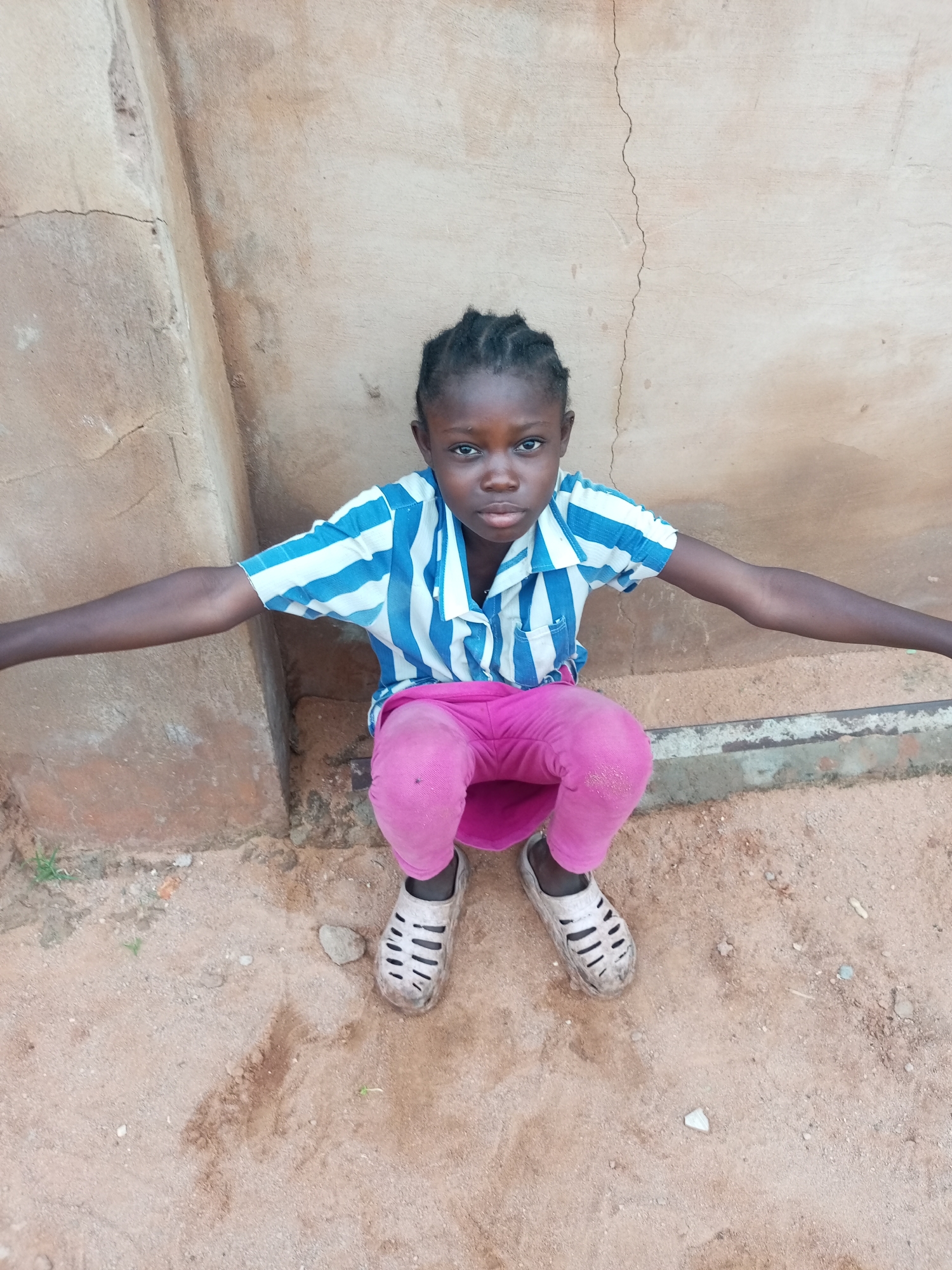 A young girl with dark skin and braided black hair kneels on sandy ground in front of a textured, tan-colored wall. She is wearing a blue and white striped short-sleeved shirt and bright pink pants. Her arms are outstretched to her sides. Her feet are clad in dirty, white clogs. The wall behind her has visible cracks and appears weathered. Small patches of green vegetation are scattered on the ground around her. The overall impression is of an outdoor setting, possibly a residential area, in Tempure, Nigeria.