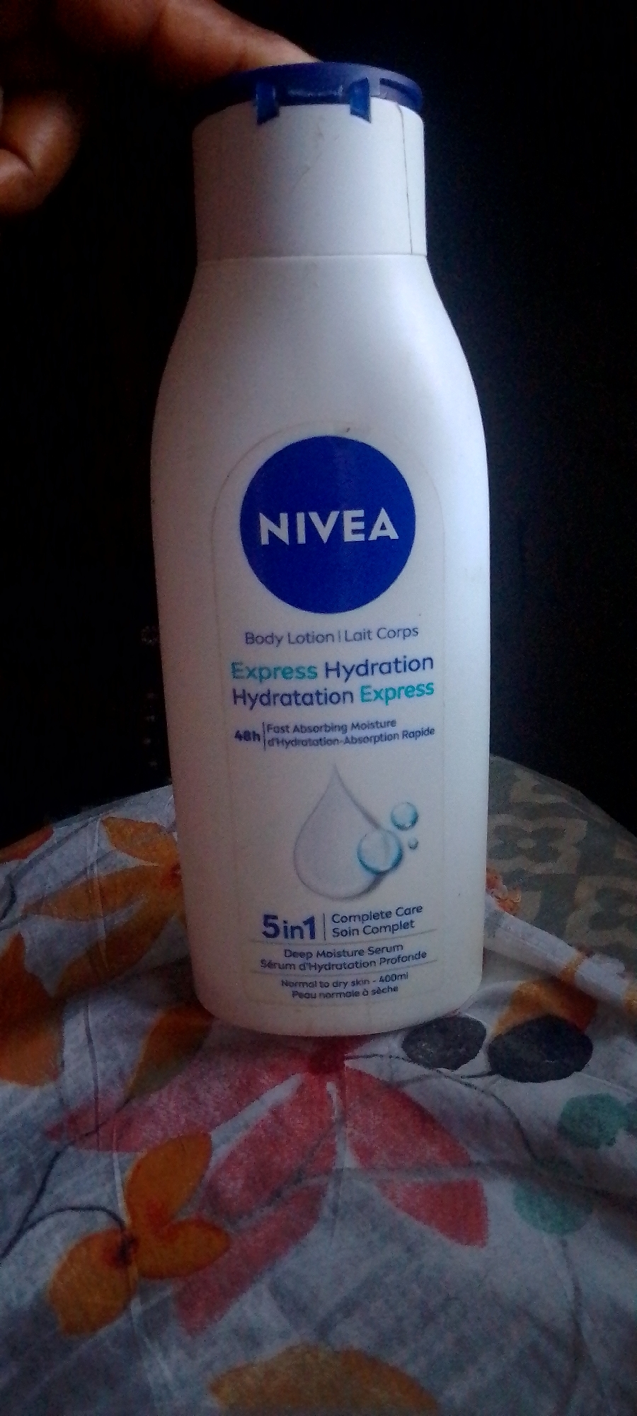 The image depicts a white Nivea "Express Hydration" body lotion bottle, positioned vertically against a dark, indistinct background. The bottle has a dark blue flip-top cap, partially obscured by the fingers and thumb of a human hand with dark skin tone, visible at the top left. The bottle's label features a central blue circle with "NIVEA" in white text. Below this, the product is identified in English and French as "Body Lotion | Lait Corps" and "Express Hydration | Hydratation Express." Additional text describes it as providing "48h Fast Absorbing Moisture | d'Hydratation-Absorption Rapide" and being "5in1 Complete Care | Soin Complet," containing "Deep Moisture Serum | Sérum d'Hydratation Profonde." A graphic of a large water droplet with smaller droplets accompanies this information. The lotion is specified for "Normal to dry skin | Peau normale à sèche" and has a volume of "400ml." The base of the bottle rests on a patterned fabric surface, displaying abstract leaf-like or floral designs in shades of orange, red, and dark grey/black on a lighter, possibly greyish-white, background. The scene is illuminated evenly, highlighting the bottle and the fabric. The location is an indoor setting; the specific city and state are unknown.