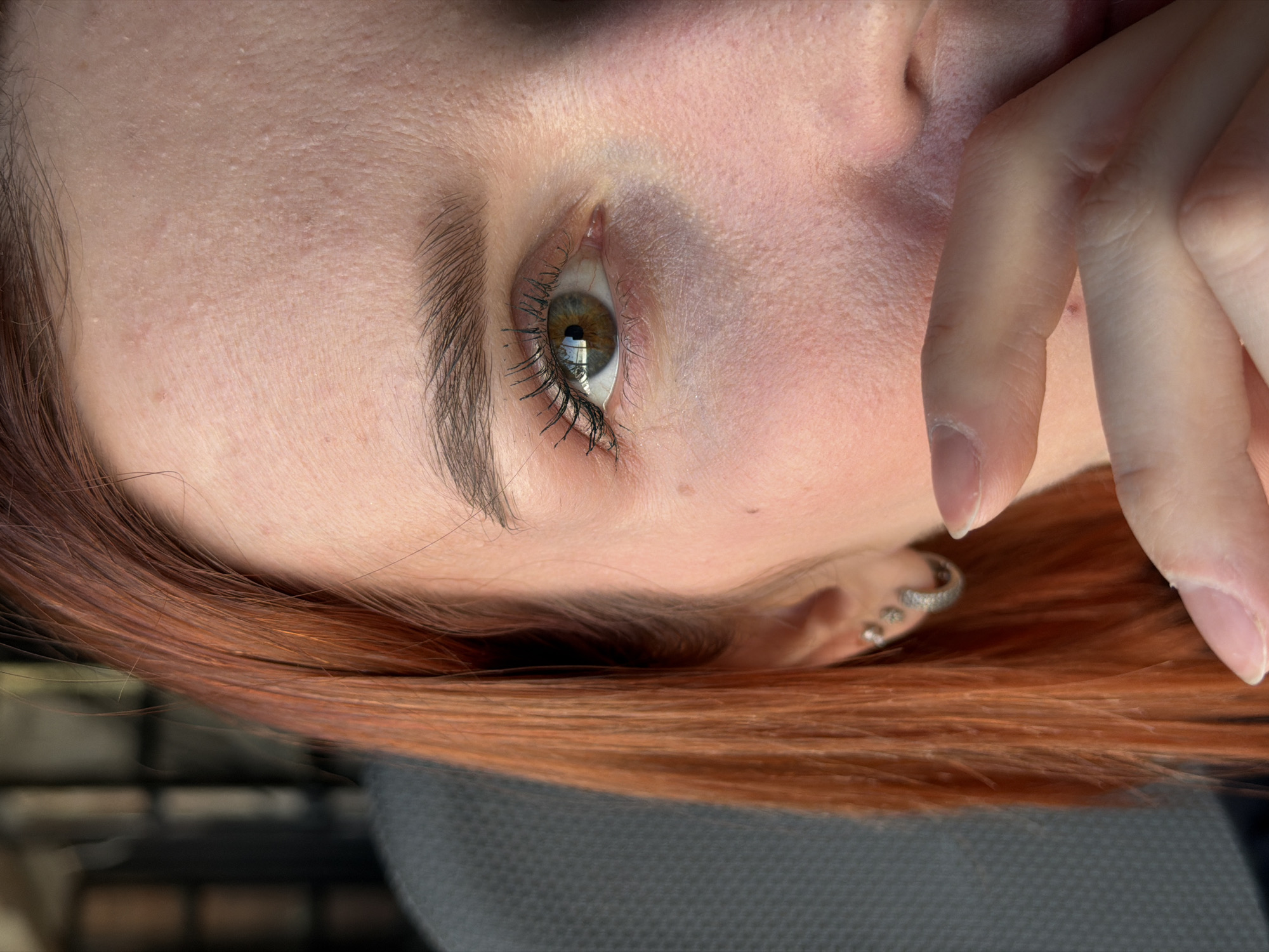 A close-up, vertical portrait captures the right side of a female subject's face. The subject has reddish-brown hair and fair skin, exhibiting visible pores and minor texture variations, including small papules on the forehead and a small nevus inferior to the right eye. The right eye features a hazel iris with discernible reflections of an external light source, and eyelashes appear darkened, consistent with mascara application. A dark brown eyebrow is present. The right earlobe is pierced in three locations: one larger silver-colored hoop earring with small stones on the lower lobe, and two smaller silver-colored stud earrings positioned superior to it. The subject's right hand covers the lower face, obscuring the mouth and lower nose, with the thumb and index finger prominent. Fingernails are natural and unpainted. The background is out of focus, presenting a dark gray, subtly textured surface on the left, consistent with an upholstered seat, and indistinct darker forms further left. The specific location within the city of Chaikovsky, Russia, is not discernible from the visual evidence.