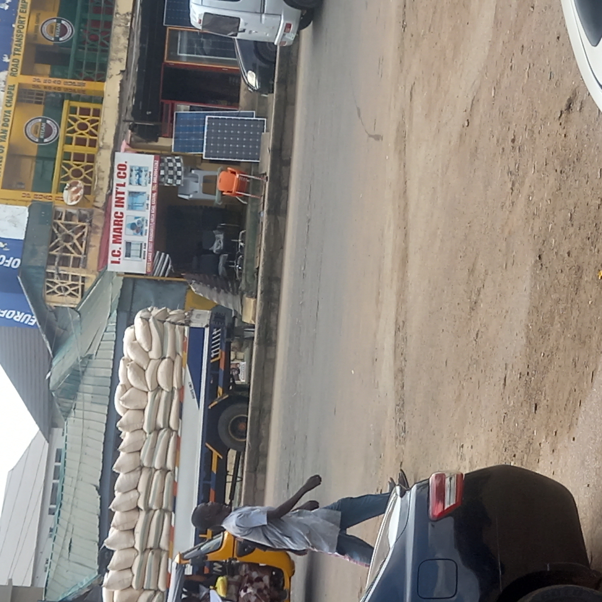 The image depicts a vibrant street scene in a commercial district, likely in Sardauna, Nigeria, under bright daylight conditions. The foreground features a dusty roadside next to a paved road. A man, dressed in a light-colored shirt and jeans, is captured in motion, walking past the rear of a dark blue car.
Further down the street, a large truck is conspicuously loaded with numerous stacks of white and striped sacks, indicating the transport of bulk goods. Adjacent to the truck, a yellow and black tricycle (likely a keke napep) is visible.
Along the left side of the street, various businesses line the road. One building, characterized by its yellow and green facade, displays a sign that reads "YAN DOYA CHAPEL ROAD TRANSPORT EMP" and features "KITEAN" logos. Nearby, a prominent white sign for "I.C. MARC INT'L CO." is visible, with smaller text underneath stating "OFFICE LAKO STREET KADUNA TEL: 08039943282." Merchandise like several solar panels (both blue and black) and various types of chairs are displayed outside these establishments. Partially visible signs include text like "EUROF" and "OFO." The environment appears bustling and dusty, typical of an active commercial street.