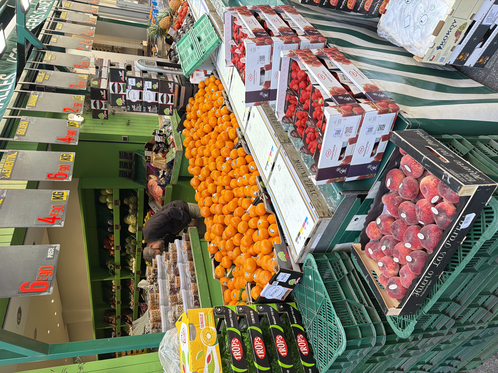The image shows an outdoor fruit and vegetable stand, likely in Paris, France, as indicated by the French language on the signs and packaging. A person is seen bending down to examine the produce. The stand is stocked with a variety of items including oranges, strawberries in boxes, peaches in a crate, pineapples, avocados, kiwis (branded as "Zespri SUNGOLD"), and fruits packaged in plastic containers. Some products are labeled with prices per kilogram or piece. The stand has a green structure with striped awning.