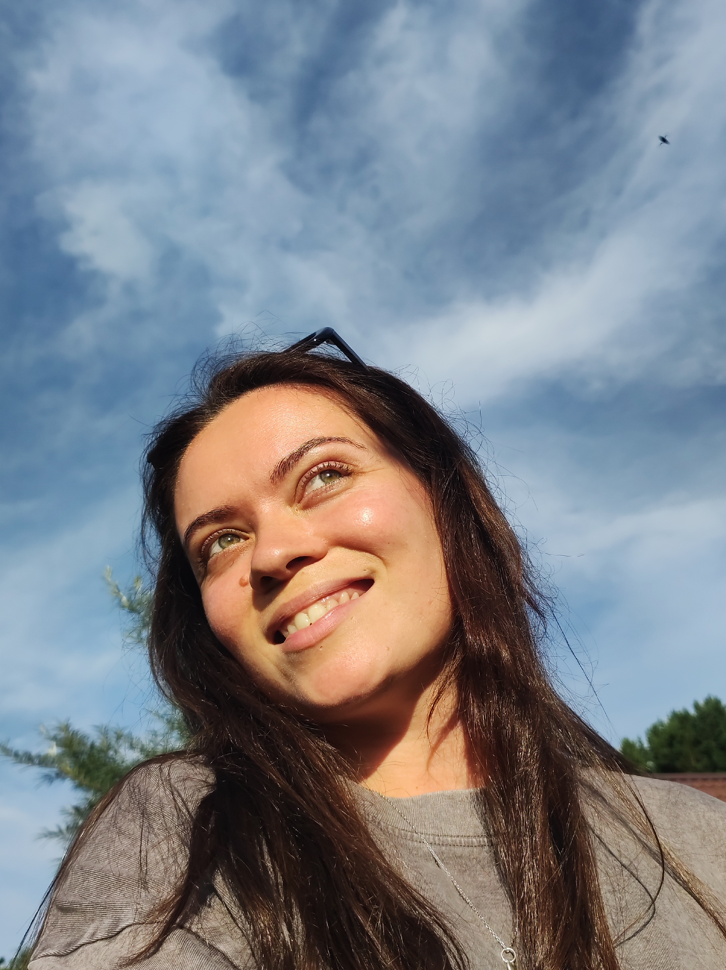 A medium shot captures a young woman with long, dark brown hair looking up towards a bright, cloudy blue sky. She is wearing a grey t-shirt and a silver necklace. Sunlight illuminates her face from the right, casting a shadow across her left cheek and the left side of her hair. Her green eyes are looking upwards, and she has a wide, beaming smile, showing her teeth. Her eyebrows are dark and well-defined, and her skin appears smooth and lightly tanned. A pair of dark-rimmed sunglasses rests on top of her head. In the background, the sky is filled with soft, white clouds, and a small, dark bird is visible in flight in the upper right portion of the frame. To the lower left of the woman, the branches of a green evergreen tree are partially visible, and to the far right, the tops of dark green trees and a brown roofline can be seen, suggesting an outdoor setting, possibly a residential area. The overall impression is one of joy and optimism under a clear sky. This scene is likely located in the city of Fokino, Russia, given the presence of evergreen trees and building styles common to the region.
